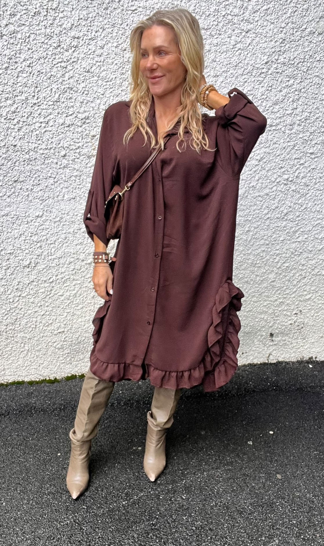 Blond kvinna står lutad mot en ljus, grovputsad vägg och ler åt sidan, med ena handen bakom nacken. Hon bär en vinröd skjortklänning med volanger, beige byxor, stövlar och liten axelväska.