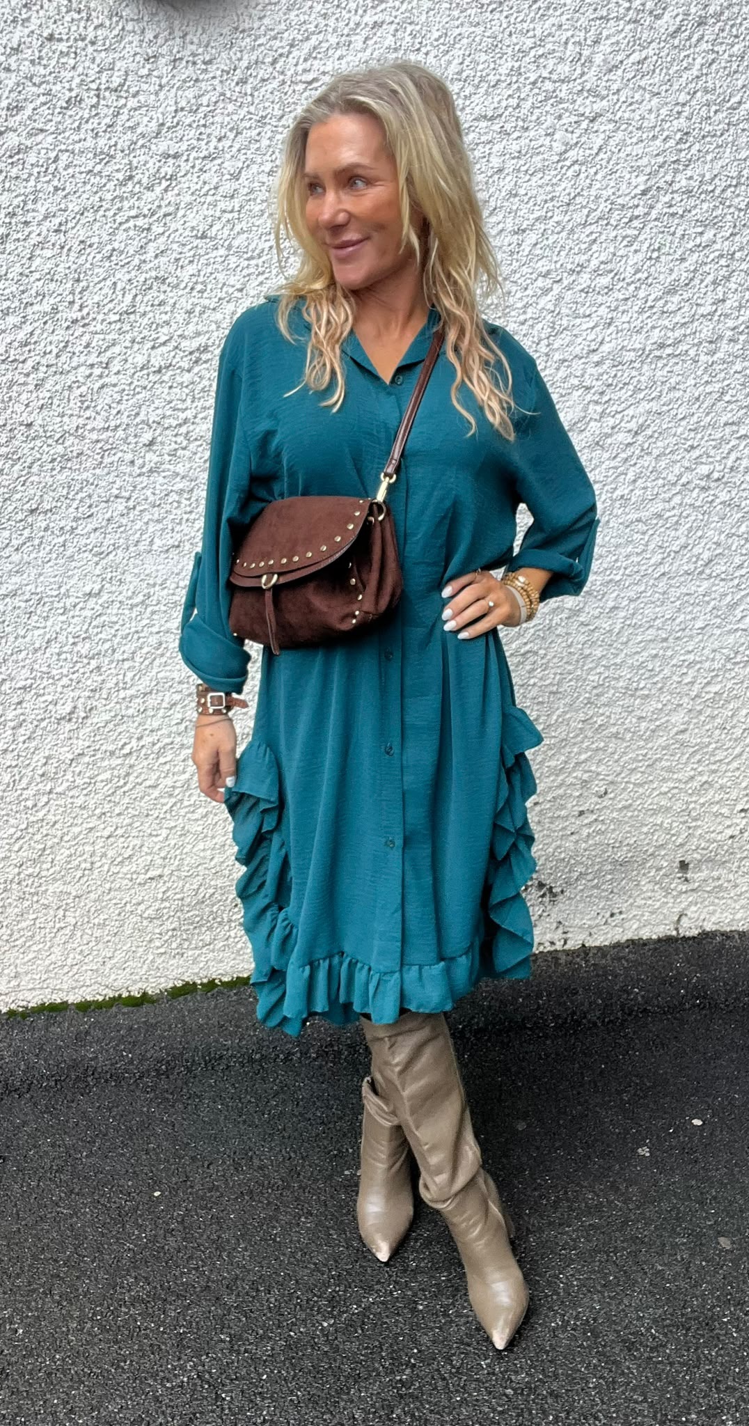 Blond kvinna står och poserar mot en vit, grovputsad vägg, ler och tittar åt sidan. Hon bär en tealgrön klänning med volanger, brun axelväska med nitar samt beige knähöga stövlar på asfalterad mark.
