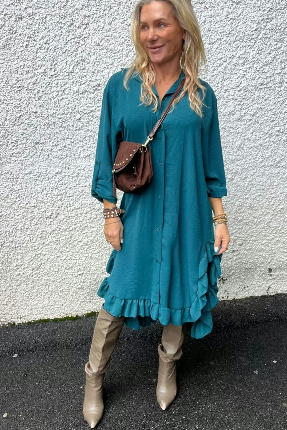 Kvinna med långt blont hår står och ler lätt, vänd åt sidan, framför en vit putsad vägg. Hon bär turkos skjortklänning med volangkant, bruna axelväska och beige knähöga stövlar på asfalt.