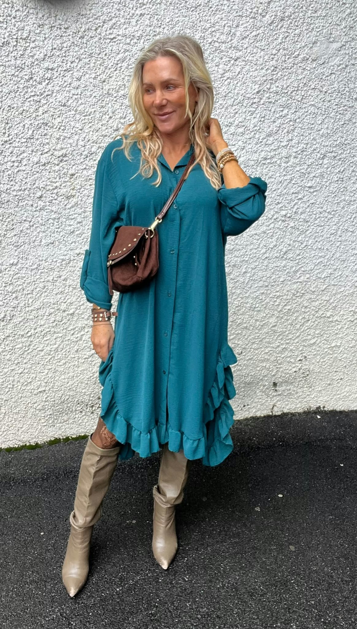 Blond kvinna står lutad mot en vit, grovputsad vägg och tittar åt sidan med ett leende. Hon bär turkos skjortklänning med volangkant, beige knähöga stövlar, armband och en brun axelväska.