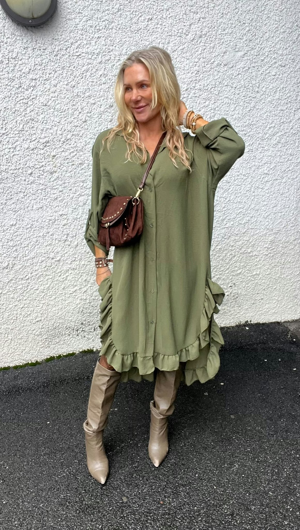 Blond kvinna står lutad mot en ljus, grovputsad vägg och ler medan hon håller ena handen i håret. Hon bär en olivgrön skjortklänning med volangkant, brun crossbody-väska, armband och beige spetsiga stövlar.