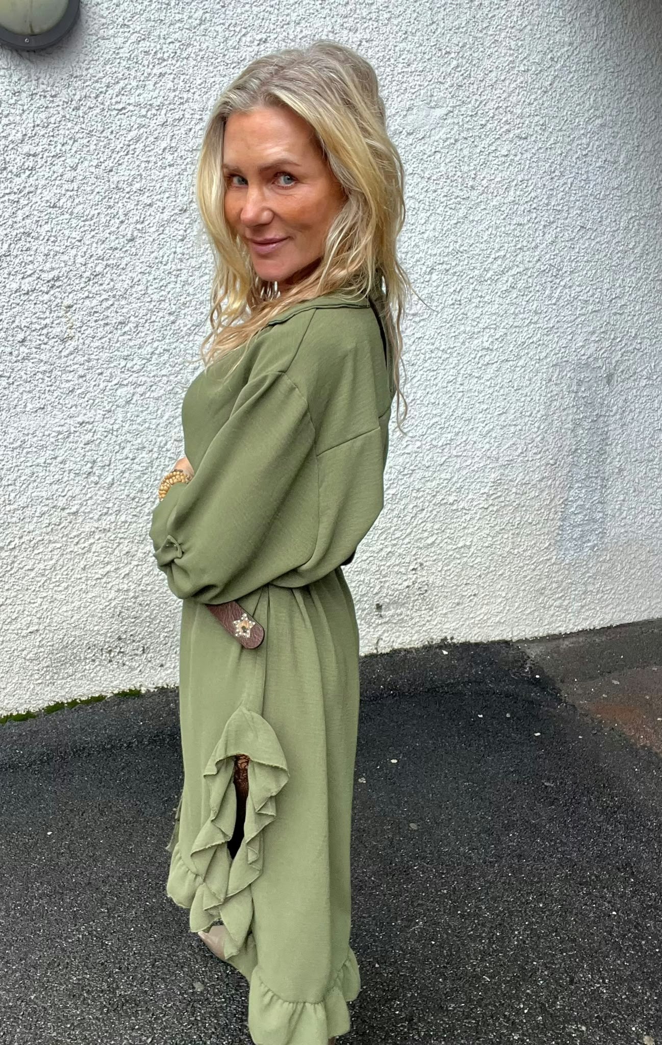 Blond kvinna i lång, olivgrön klänning står i profil med armarna i kors och vänder blicken mot kameran. Hon står utomhus på asfalt framför en vit, grovputsad vägg; ett runt väggljus syns uppe till vänster.