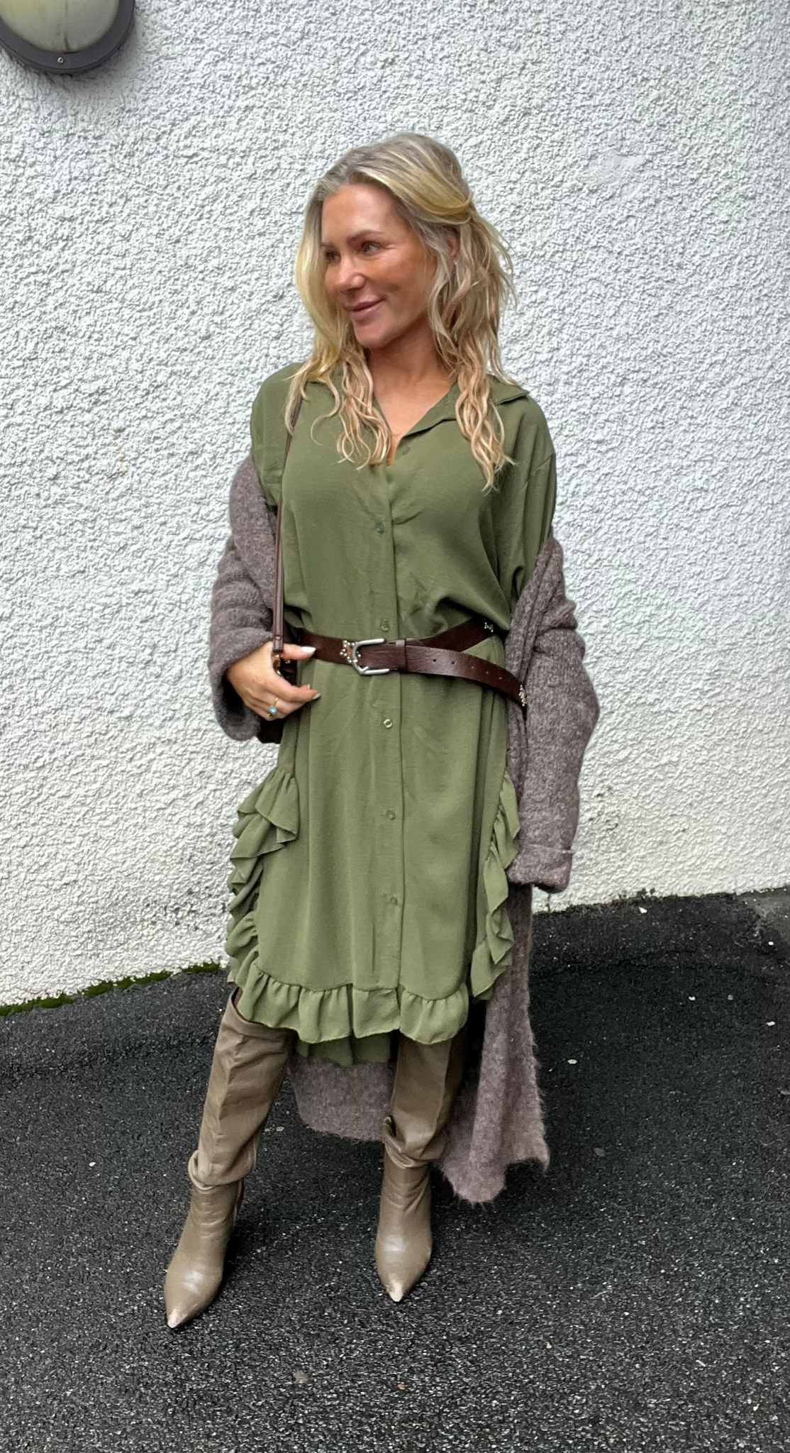 Kvinna står mot en vit, grovputsad vägg och tittar åt sidan med ett svagt leende. Hon bär grön skjortklänning med volanger och brunt bälte, lång brun kofta samt beige stövlar, och håller en axelrem.