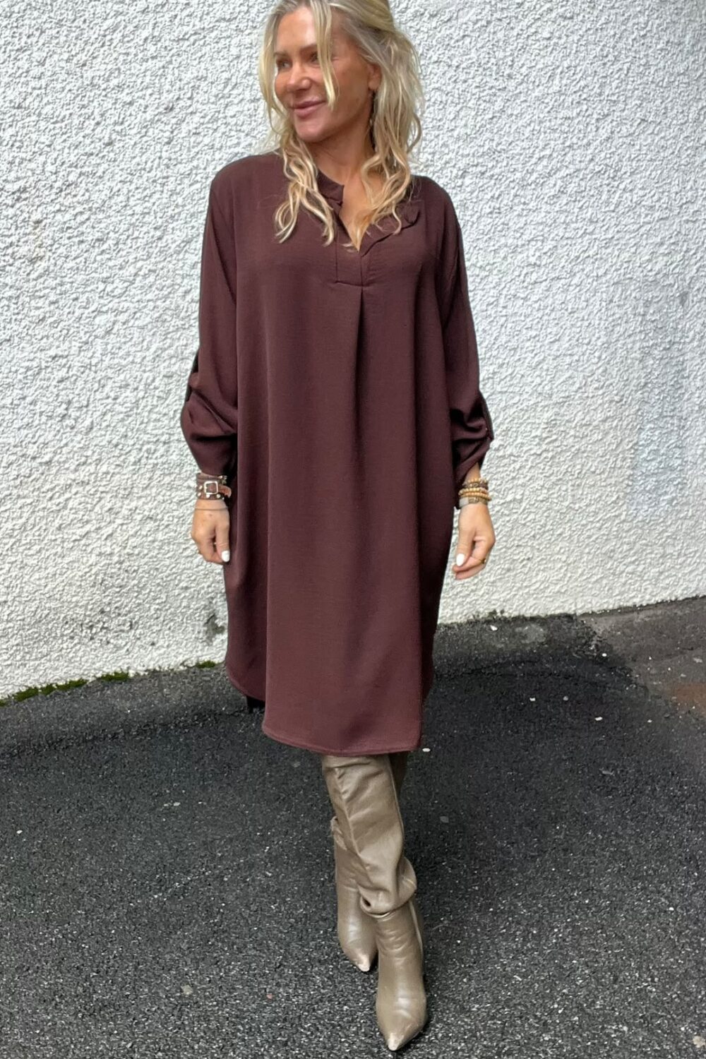Blond kvinna står avslappnat mot en vit, grovputsad vägg och tittar åt sidan. Hon bär en brun, knälång klänning med långa ärmar, beige knähöga stövlar och flera armband, på asfalt utomhus.