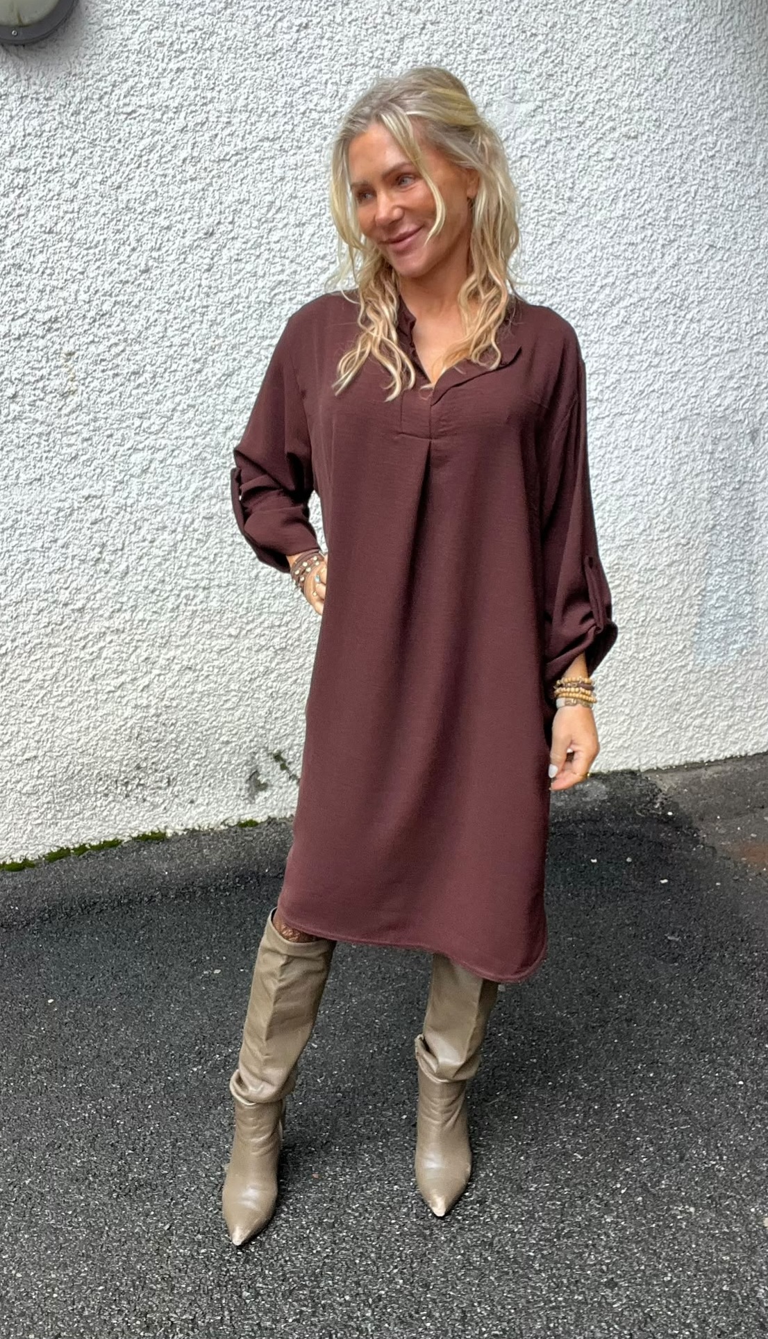 Leende kvinna med långt blont hår står avslappnat mot en ljus, grov vägg och tittar åt sidan. Hon bär en vinröd, lång skjortklänning, beige stövlar, smala byxor och armband på handlederna.