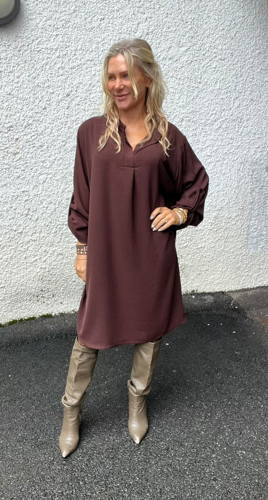 Blond kvinna står utomhus mot en vit, grovputsad vägg och ler åt sidan. Hon bär en lång, vinröd tunika med trekvartsärm, beige byxor och spetsiga beige stövlar samt flera armband.