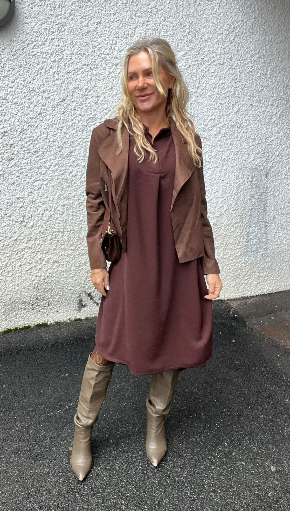 Blond kvinna står utomhus mot en ljus, grovputsad vägg och ler åt sidan. Hon bär en vinröd klänning med brun jacka, liten svart axelväska och beige knähöga stövlar på mörk asfalt.