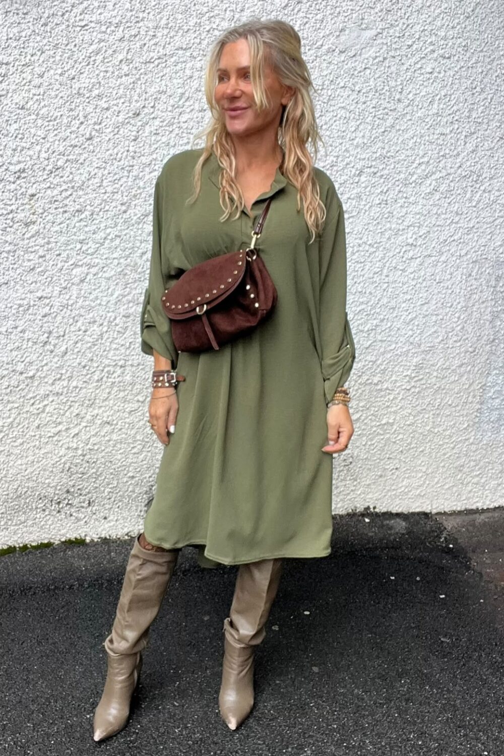 Kvinna står och poserar mot en vit, grovputsad vägg och ler åt sidan. Hon bär en olivgrön, lång skjortklänning, beige knähöga stövlar och en brun axelväska med nitar över bröstet.