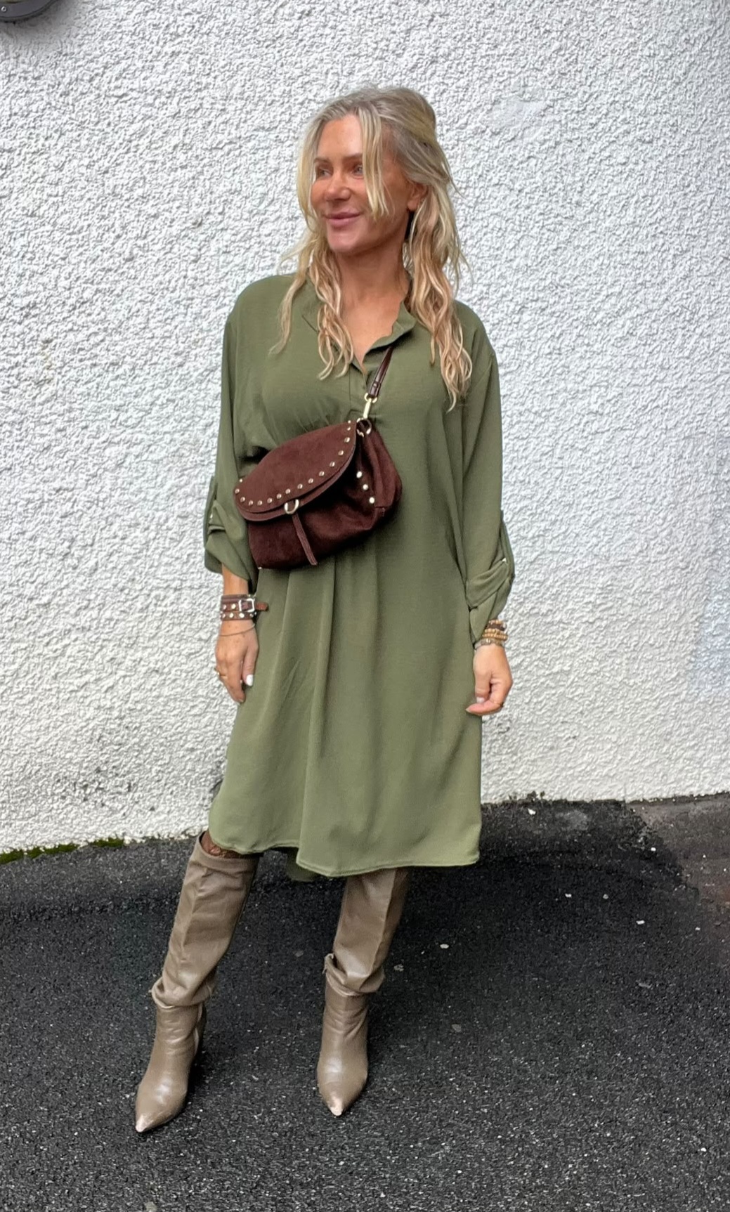Kvinna står utomhus mot en vit, grovputsad vägg och tittar åt sidan med ett leende. Hon bär en olivgrön skjortklänning, beige knähöga stövlar och en brun axelväska med nitar.