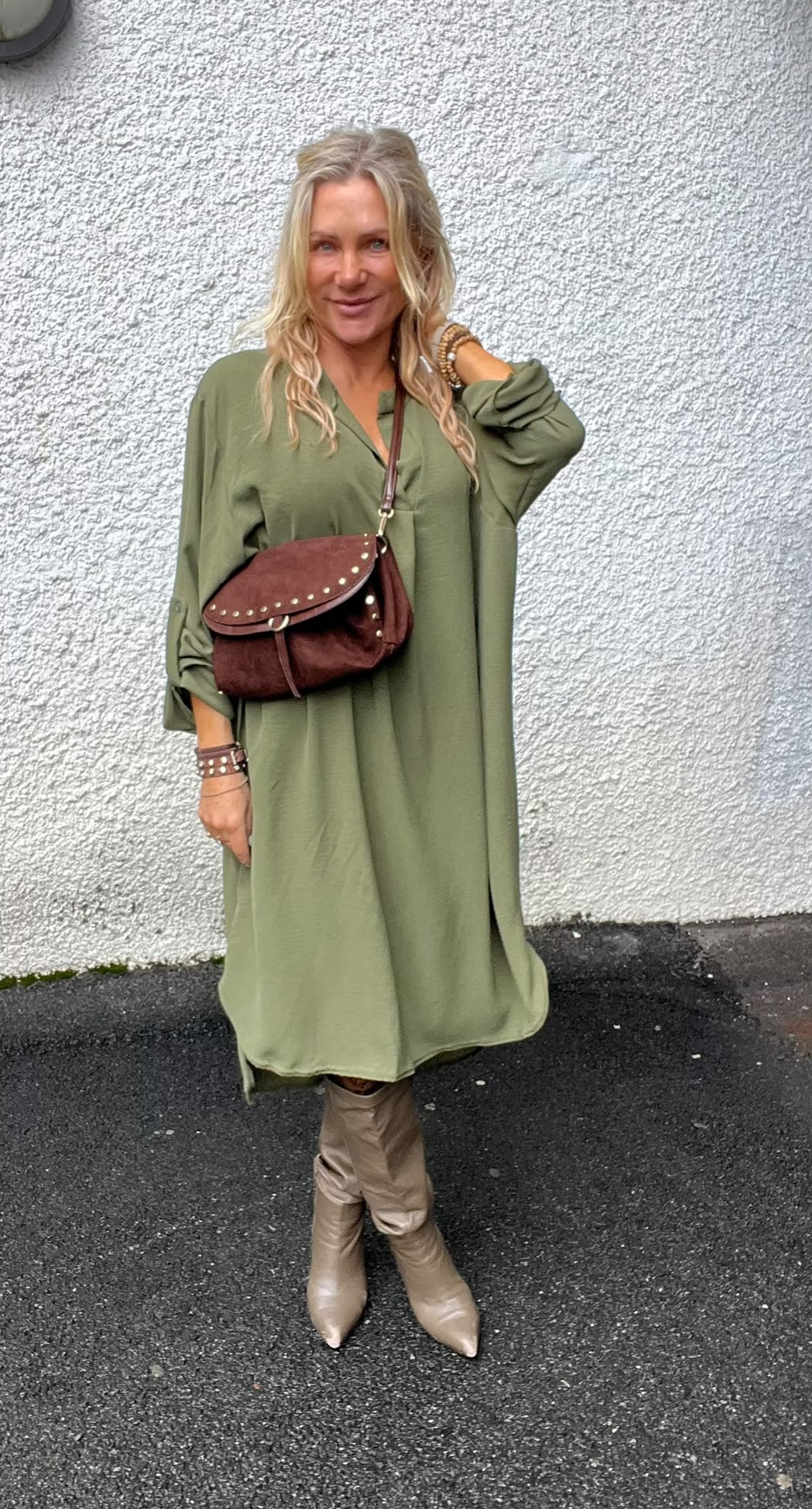 Blond kvinna står och ler mot kameran med ena handen vid håret, klädd i lång olivgrön klänning. Hon bär brun axelväska med nitar, armband och beige knähöga stövlar framför en vit putsad vägg.