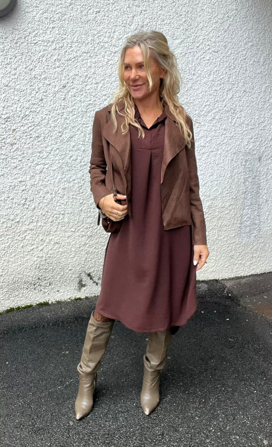 Blond kvinna står utomhus mot en ljus, grovputsad vägg och ler åt sidan. Hon bär vinröd klänning, brun jacka och knähöga beige stövlar, och håller en liten brun axelväska.