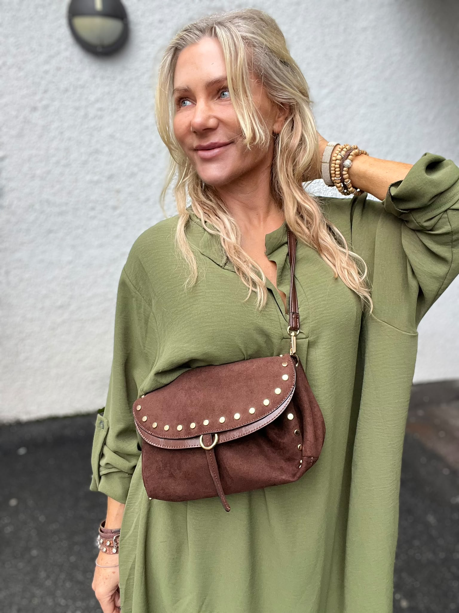 Blond kvinna i grön klänning ler och tittar åt sidan medan hon håller ena handen bakom nacken. Hon bär armband och en brun nitarprydd crossbodyväska över bröstet, framför en ljus vägg utomhus.