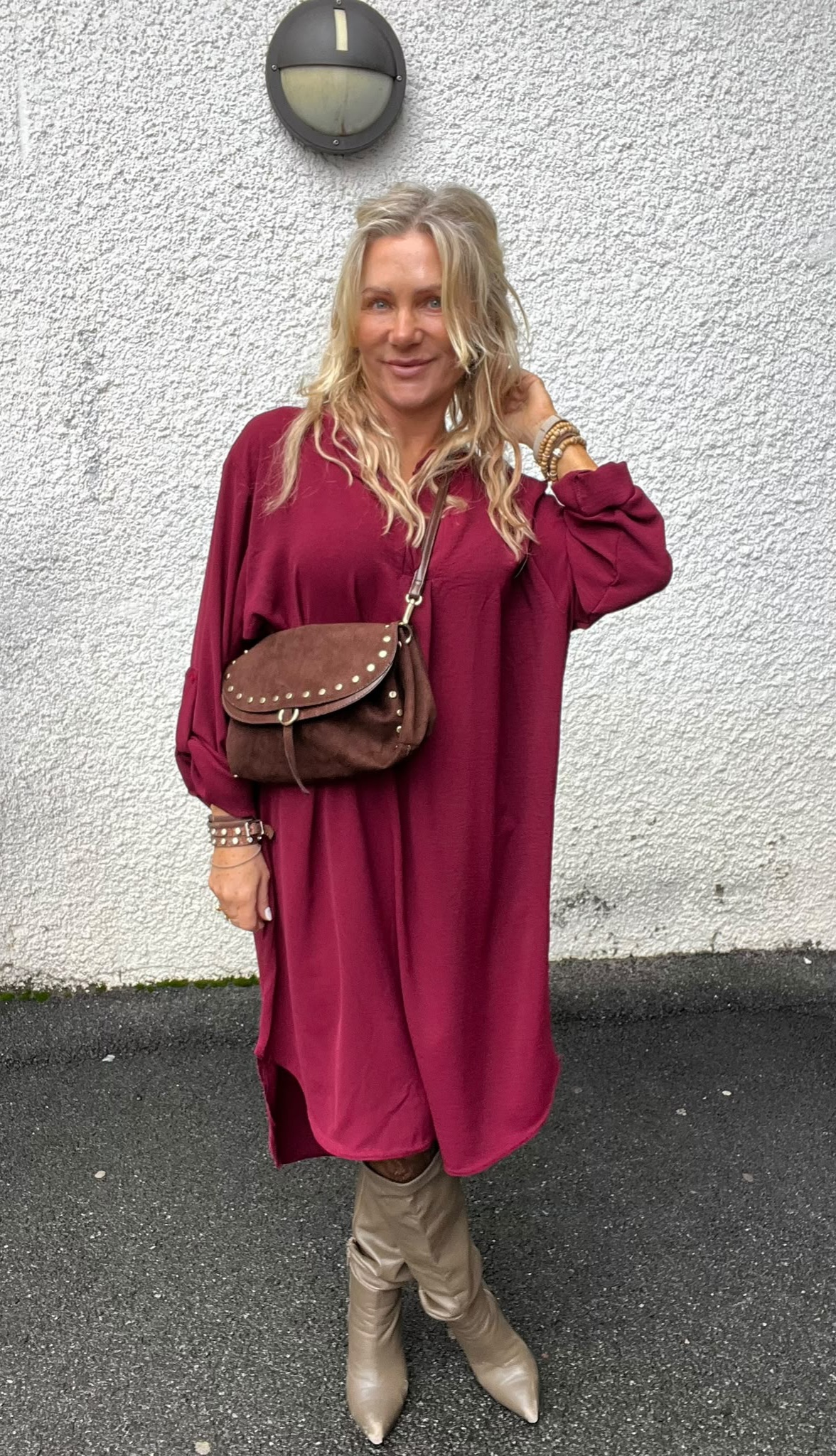 Blond kvinna står och ler mot kameran, med ena handen vid håret. Hon bär en vinröd lång klänning, ljusa knähöga stövlar och brun axelväska med nitar, framför en vitputsad vägg med rund vägglampa.