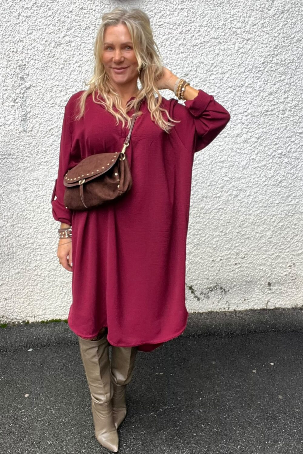Blond kvinna står och ler mot kameran med ena handen i håret, klädd i vinröd långärmad klänning. Hon bär brun axelväska med nitar och beige knähöga stövlar, framför en vit putsad vägg.