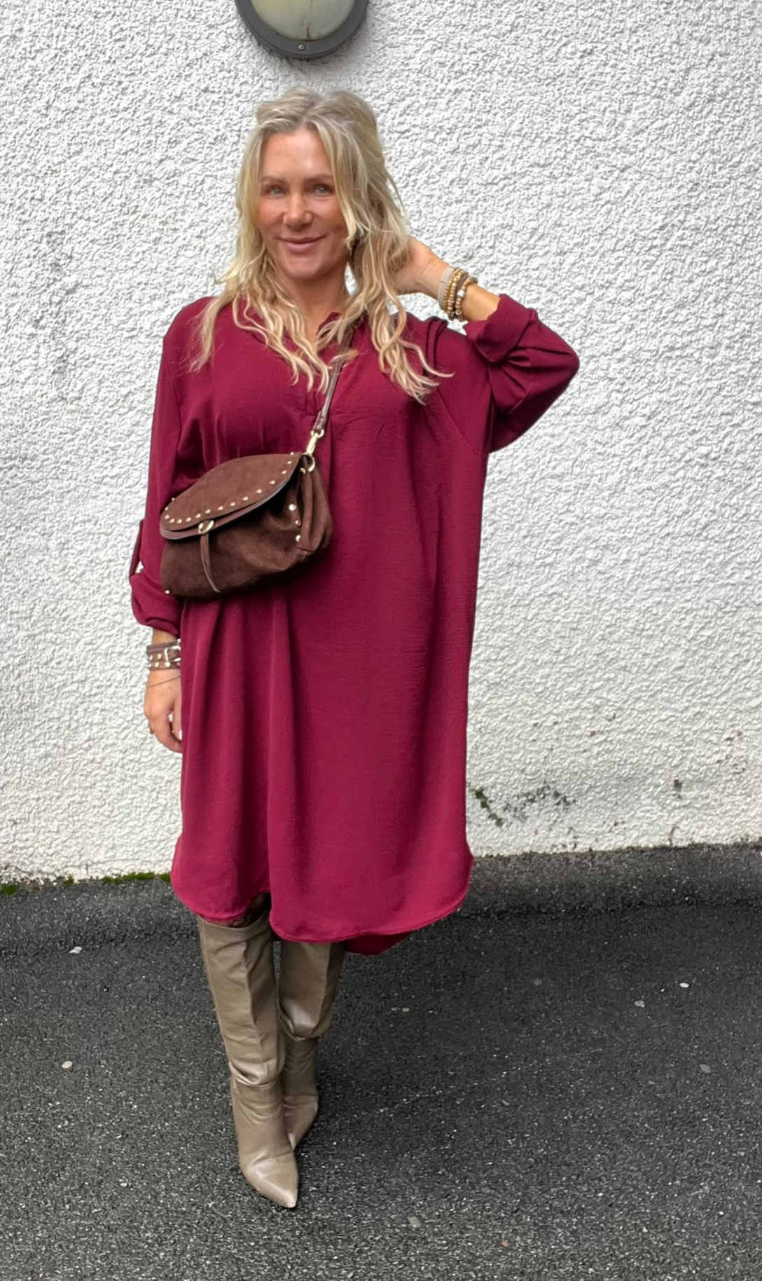 Blond kvinna står och ler mot kameran med ena handen i håret, klädd i vinröd långärmad klänning. Hon bär brun axelväska med nitar och beige knähöga stövlar, framför en vit putsad vägg.