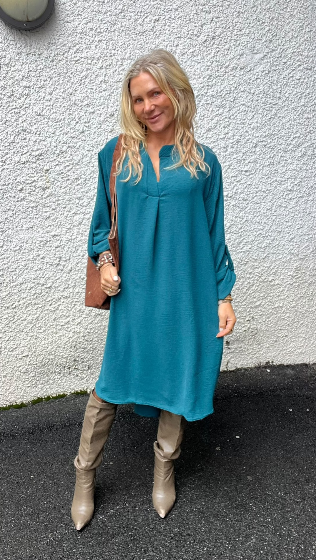 Blond kvinna står och ler mot kameran framför en vit, grovputsad vägg. Hon bär en turkos, knälång klänning, beige knähöga stövlar och en brun axelväska, med armband på handlederna.
