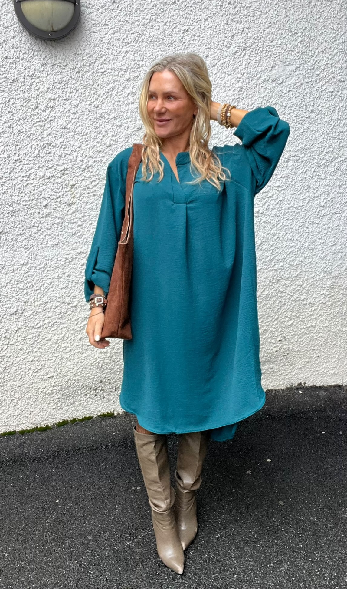 Kvinna står utomhus mot en ljus, grov vägg och ler medan hon ser åt sidan, med ena handen i håret. Hon bär en turkos klänning, höga beige stövlar och brun axelväska samt armband.