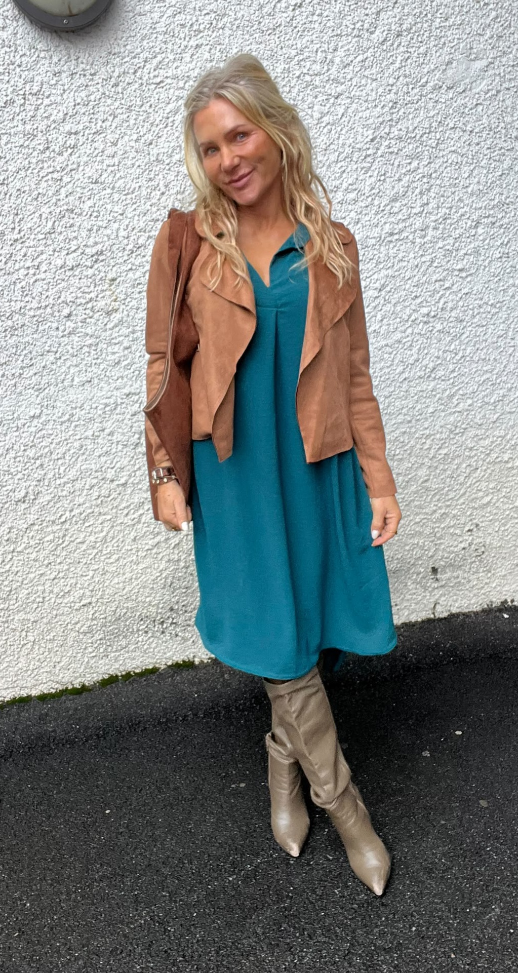 Blond kvinna står och ler mot kameran framför en vit, grovputsad vägg utomhus. Hon bär turkos klänning, ljusbrun mockajacka, höga beige stövlar och en brun axelväska, med benen lätt korsade.