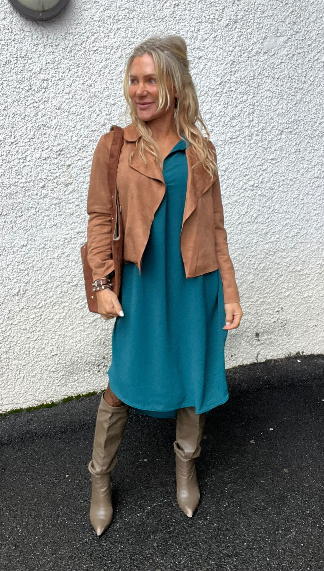 Blond kvinna står utomhus mot en vit, grovputsad vägg och ler medan hon tittar åt sidan. Hon bär turkos klänning med brun mockajacka, ljusa knähöga stövlar och brun axelväska.