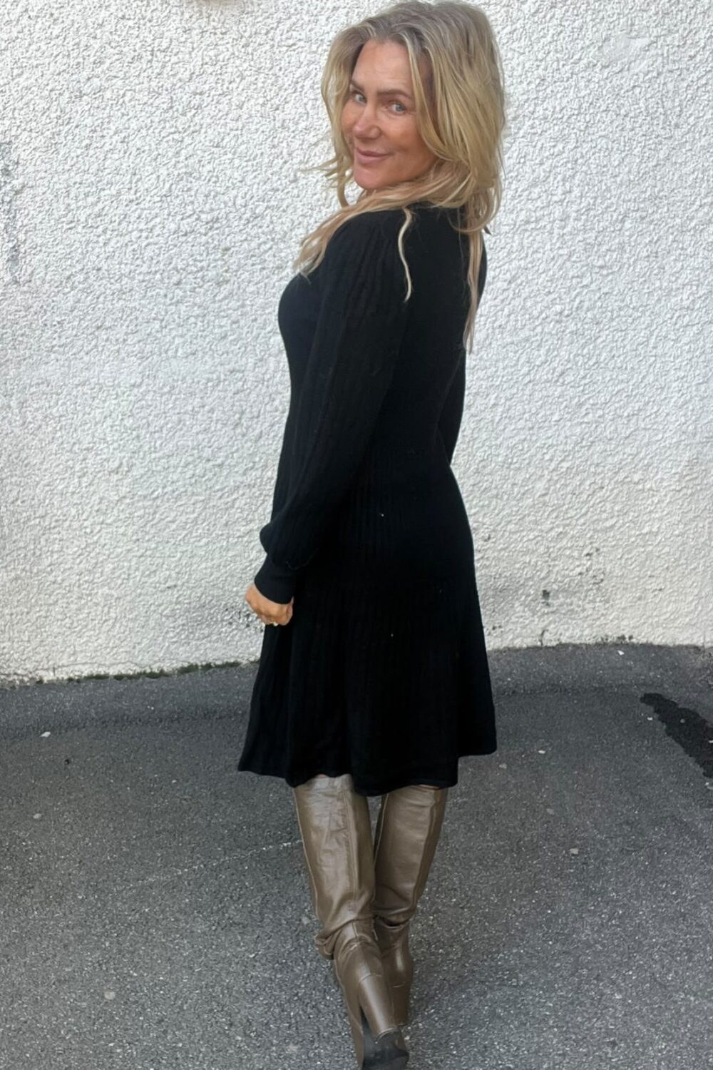 Kvinna med långt blont hår står i profil och vänder sig mot kameran med ett leende. Hon bär en svart klänning och höga, ljusbruna stövlar, och står på asfalt framför en vit putsad vägg med vägglampa.