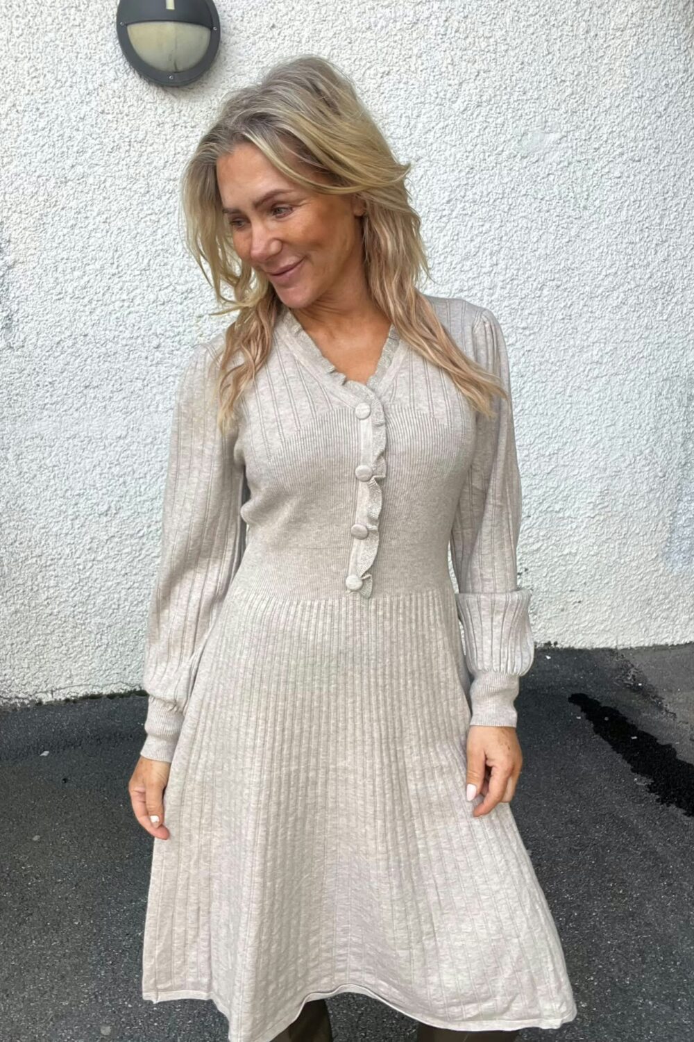 Blond kvinna står utomhus och tittar ned åt sidan med ett svagt leende, klädd i beige ribbstickad klänning med knappar framtill. Bakom henne syns vit putsad vägg med rund vägglampa och mörk asfalt.