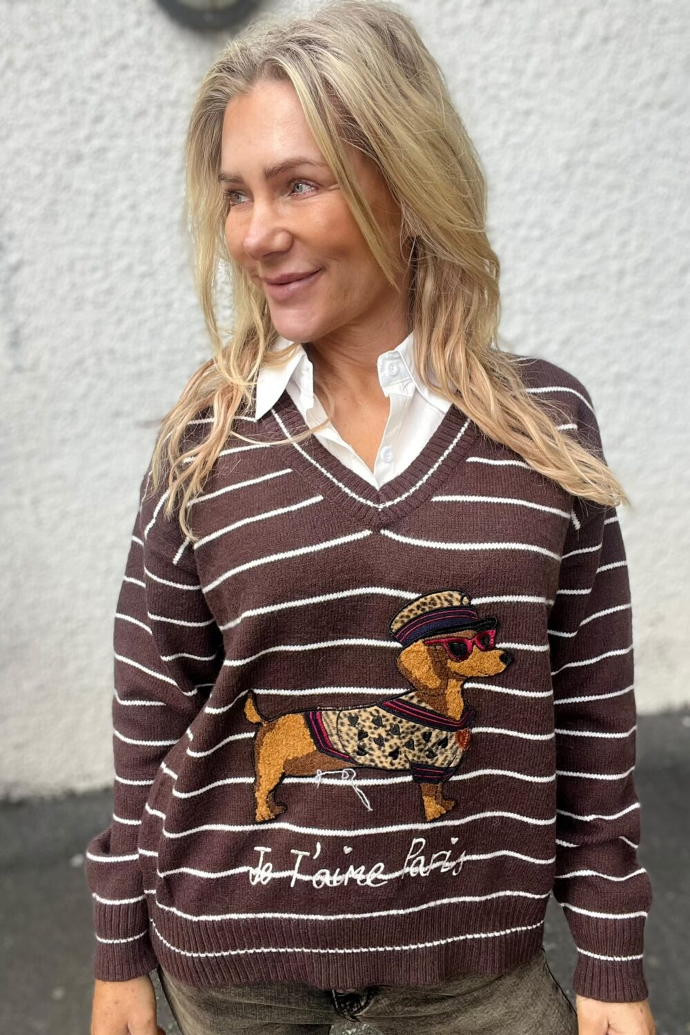 Blond kvinna står utomhus och ler medan hon tittar åt sidan, framför en ljus vägg. Hon bär vit skjorta under brunrandig tröja med broderad hund i solglasögon och texten ”Je t’aime Paris”.