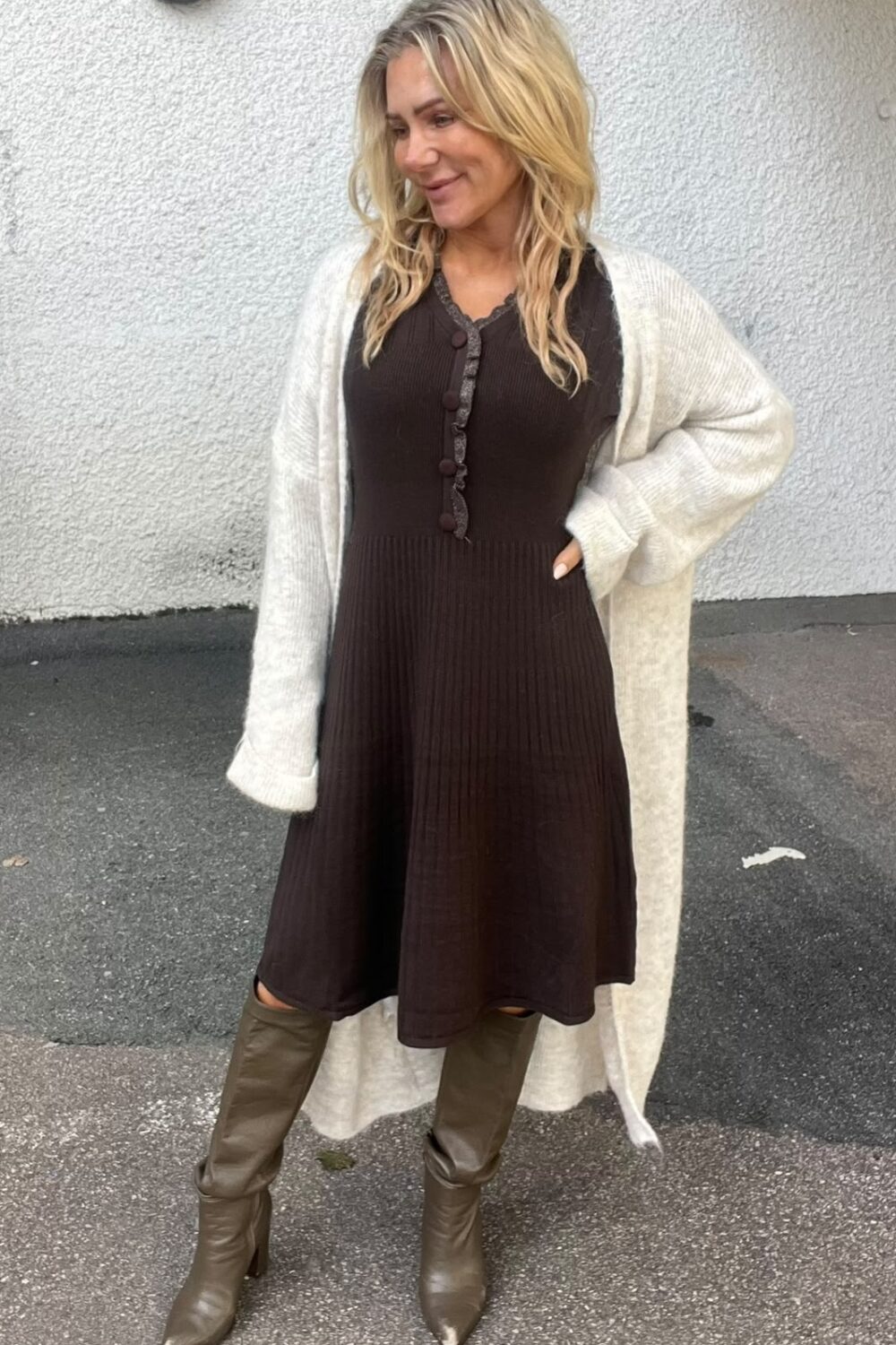 Blond kvinna står utomhus mot en ljus putsad vägg och ler åt sidan, med ena handen i fickan. Hon bär brun stickad klänning med knappar, lång ljus kofta och knähöga stövlar.