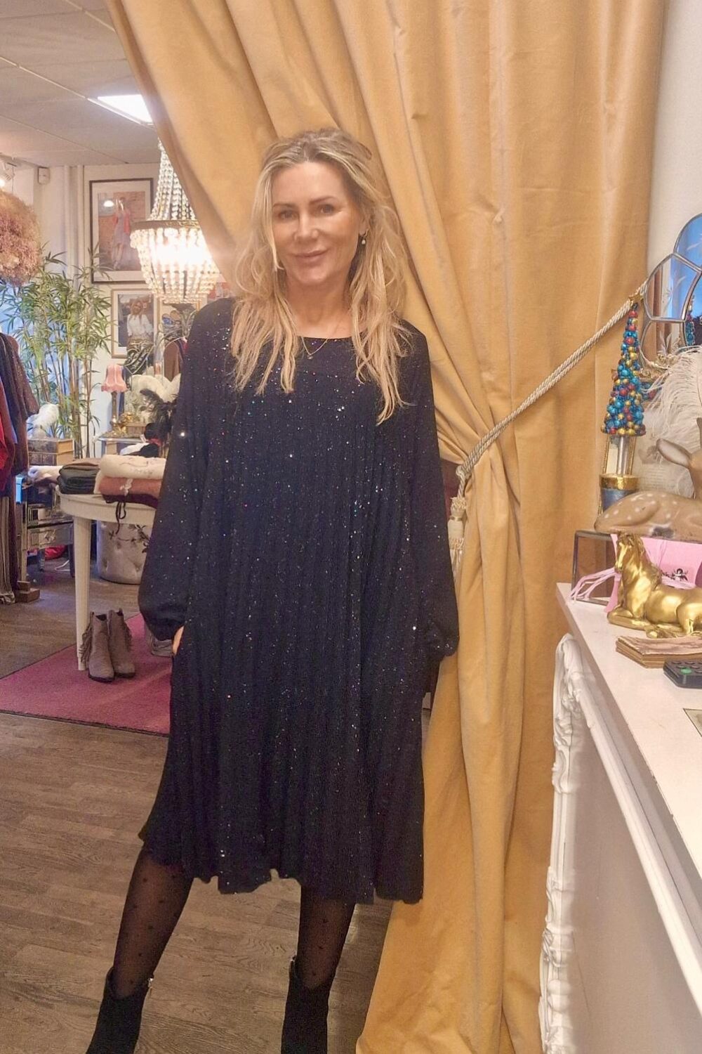 Kvinna i glittrig mörk klänning står och poserar med händerna i fickorna framför ett guldbeige draperi i en butik, omgiven av lampor, kläder, skor och dekorativa föremål på bord och hyllor.