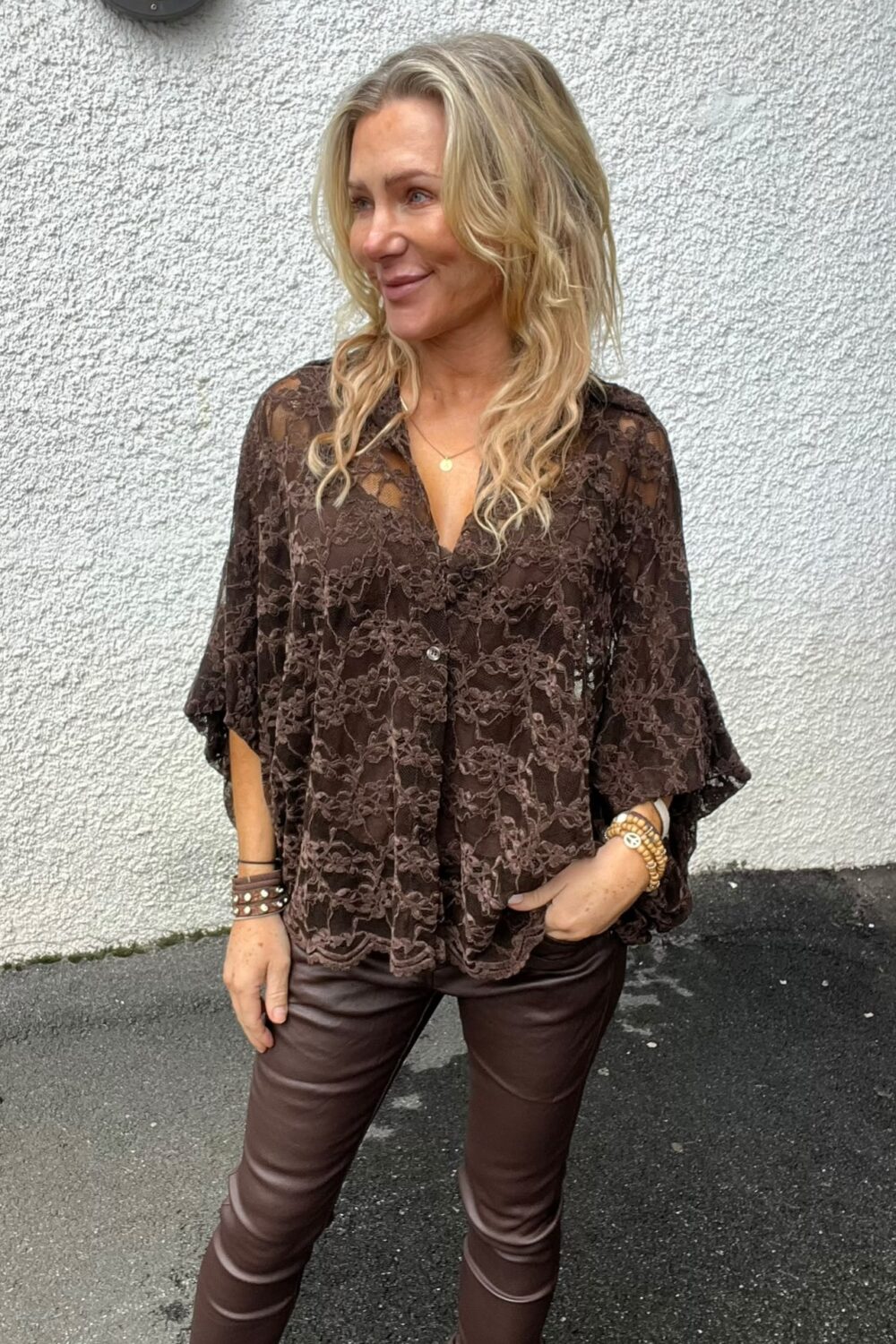 Blond kvinna står utomhus framför en vit, grovputsad vägg och ler medan hon tittar åt sidan. Hon bär en brun spetsblus, bruna skinnbyxor och armband, med ena handen i fickan.