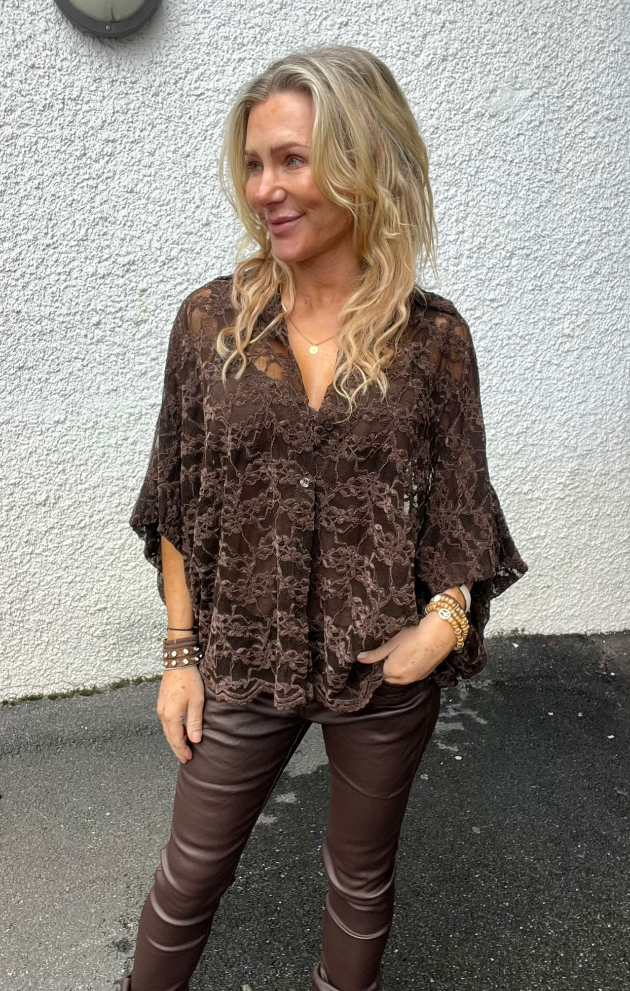 Blond kvinna står utomhus framför en vit, grovputsad vägg och ler medan hon tittar åt sidan. Hon bär en brun spetsblus, bruna skinnbyxor och armband, med ena handen i fickan.