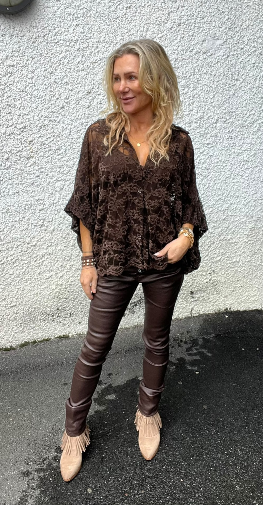 Blond kvinna står utomhus mot en ljus, grovputsad vägg och tittar åt sidan med ett leende. Hon bär en brun spetsblus, mörkbruna skinnbyxor och beige boots med fransar samt armband och halsband.