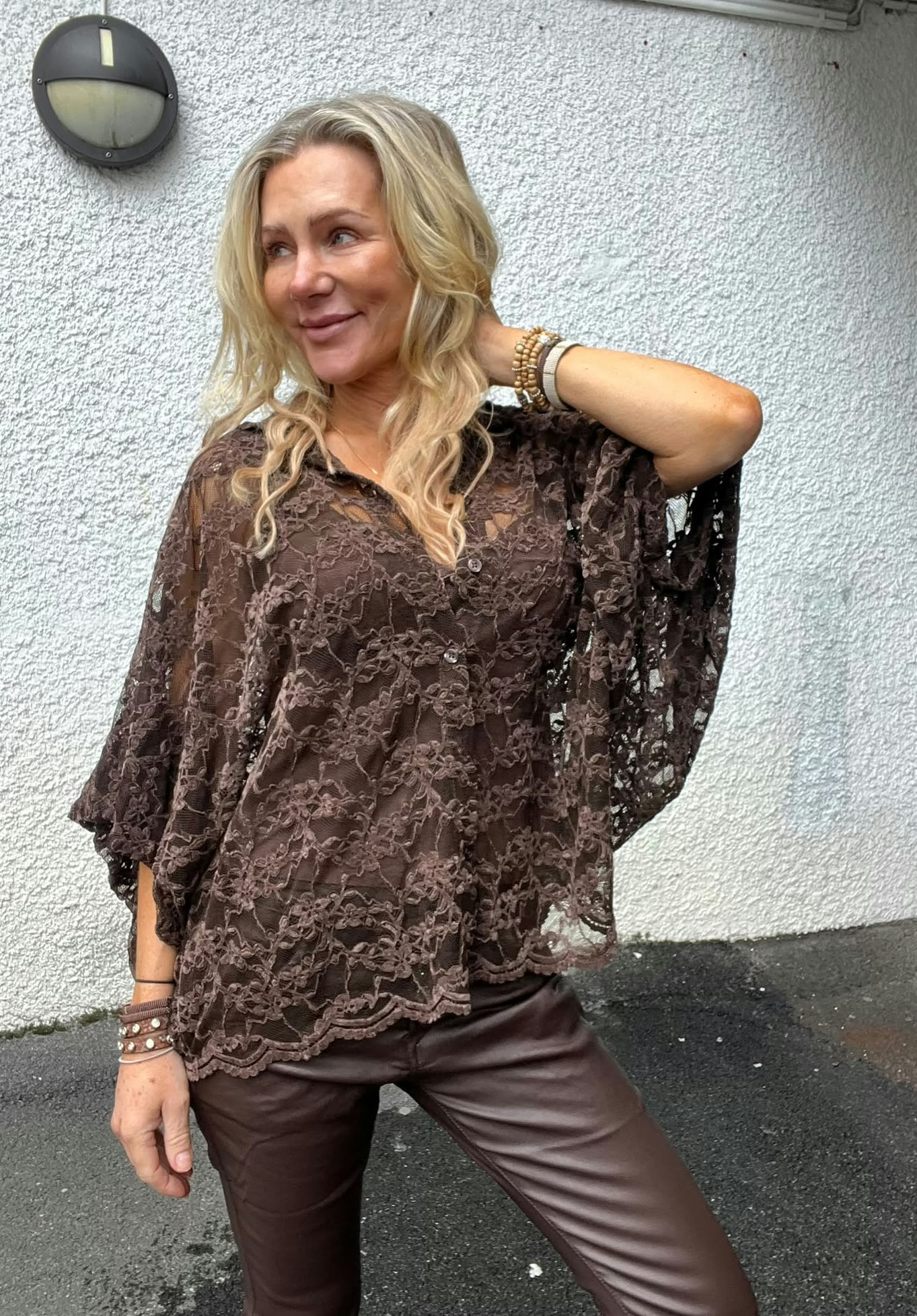 Blond kvinna poserar utomhus med ena handen bakom nacken och blicken åt sidan, framför en vit putsad vägg med rund vägglampa. Hon bär brun spetsblus med vida ärmar, bruna byxor och armband.