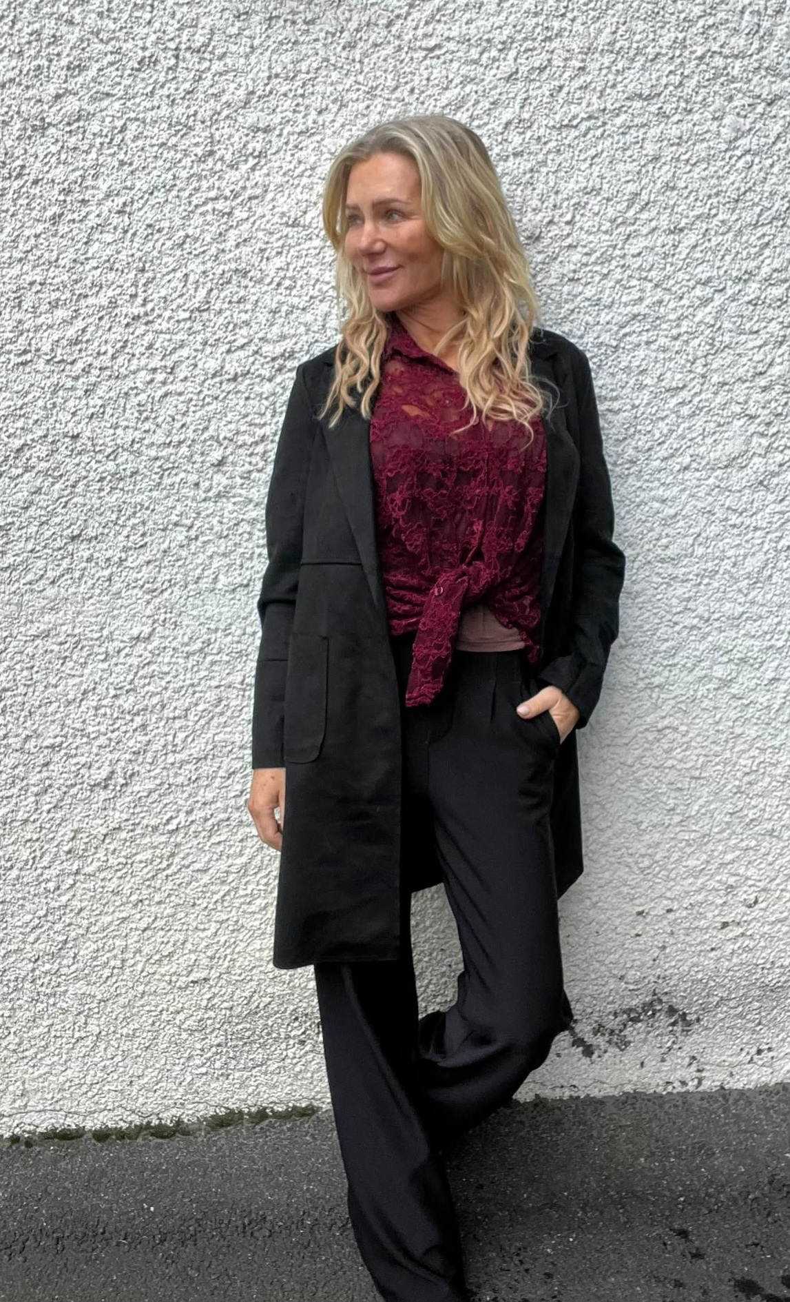 Blond kvinna står lutad mot en ljus, grovputsad vägg och ser åt sidan med ett svagt leende. Hon bär lång svart kappa, svarta byxor och vinröd spetsblus, ena handen i fickan.