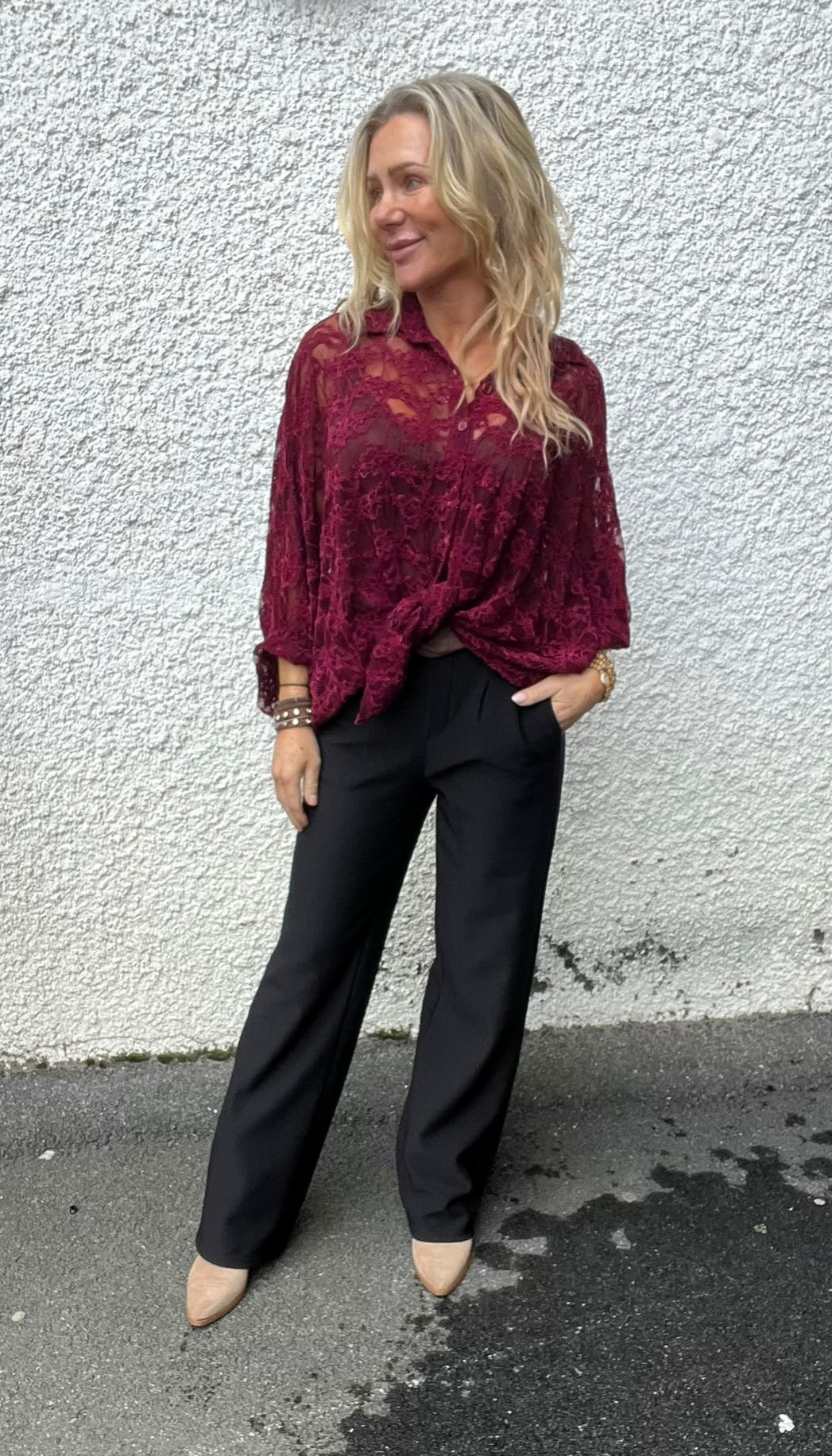 Blond kvinna står avslappnat med ena handen i fickan och tittar åt sidan. Hon bär vinröd spetsblus, svarta byxor och beige skor, med armband på handleden, mot en ljus, grovputsad vägg utomhus.