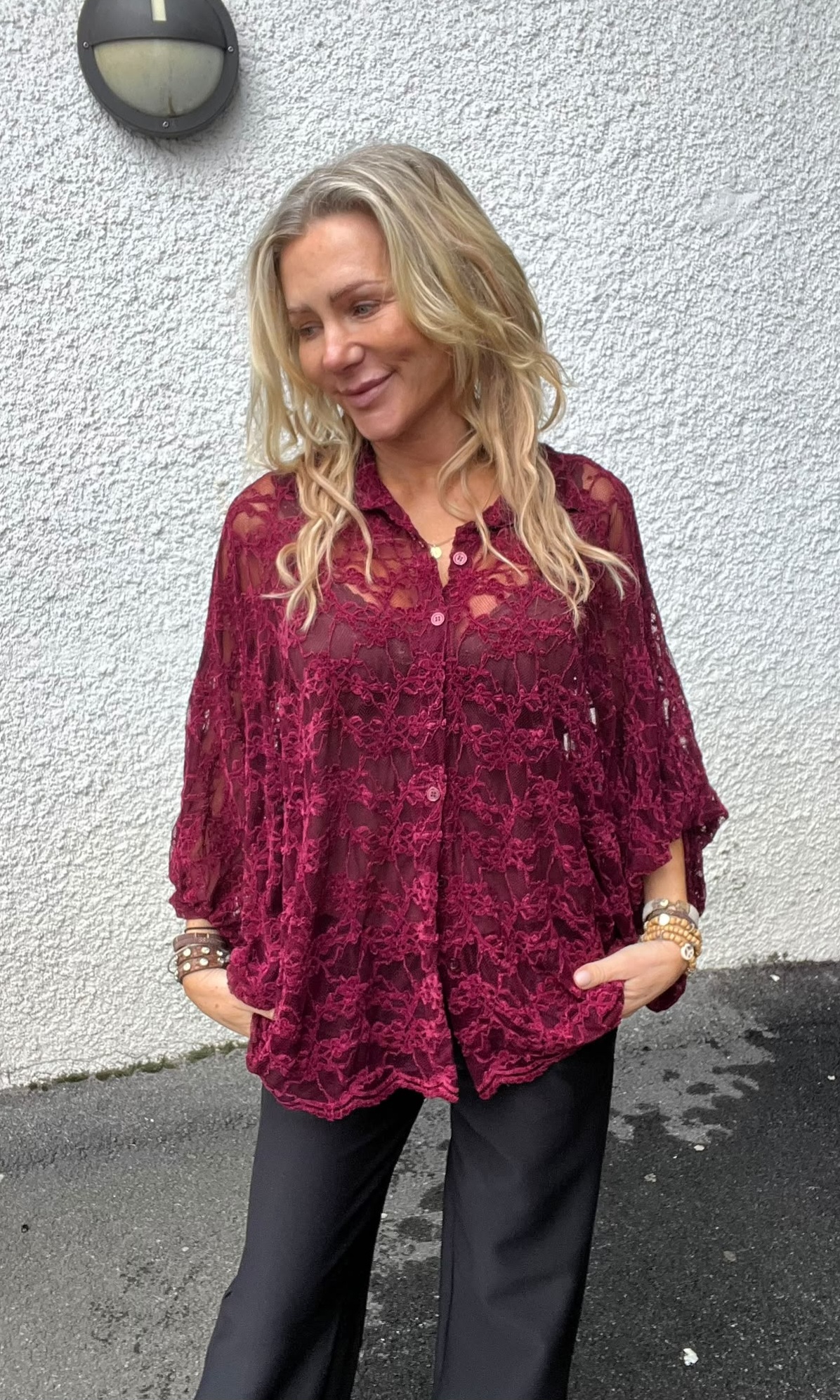 Blond kvinna står utomhus med händerna i fickorna och ler medan hon tittar åt sidan. Hon bär en vinröd spetsblus med knappar och svarta byxor, mot en vit putsad vägg med vägglampa.