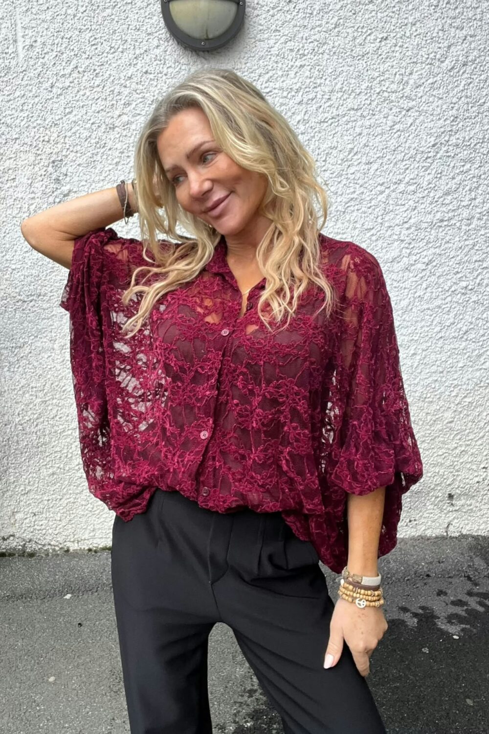 Blond kvinna poserar avslappnat med ena handen bakom huvudet och ler svagt. Hon bär en vinröd spetsblus med vida ärmar och svarta byxor, står mot en vit grovputsad vägg med rund utelampa ovanför.