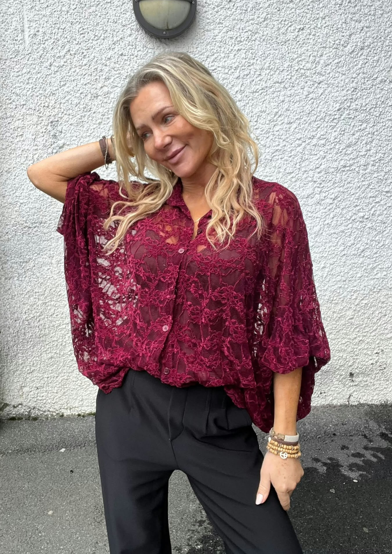 Blond kvinna poserar avslappnat med ena handen bakom huvudet och ler svagt. Hon bär en vinröd spetsblus med vida ärmar och svarta byxor, står mot en vit grovputsad vägg med rund utelampa ovanför.