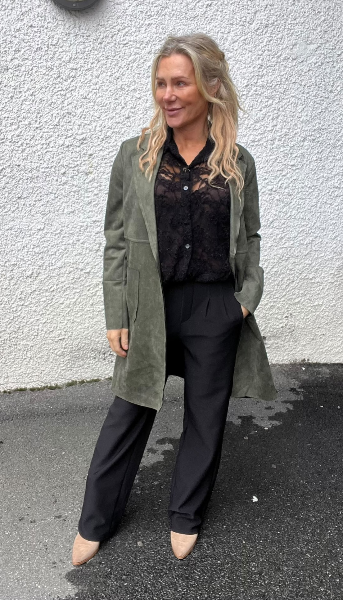 Kvinna står utomhus mot en vitputsad vägg och ler, med ena handen i fickan. Hon bär lång olivgrön kappa, svart spetsblus och svarta byxor, samt beige skor på våt asfalt.