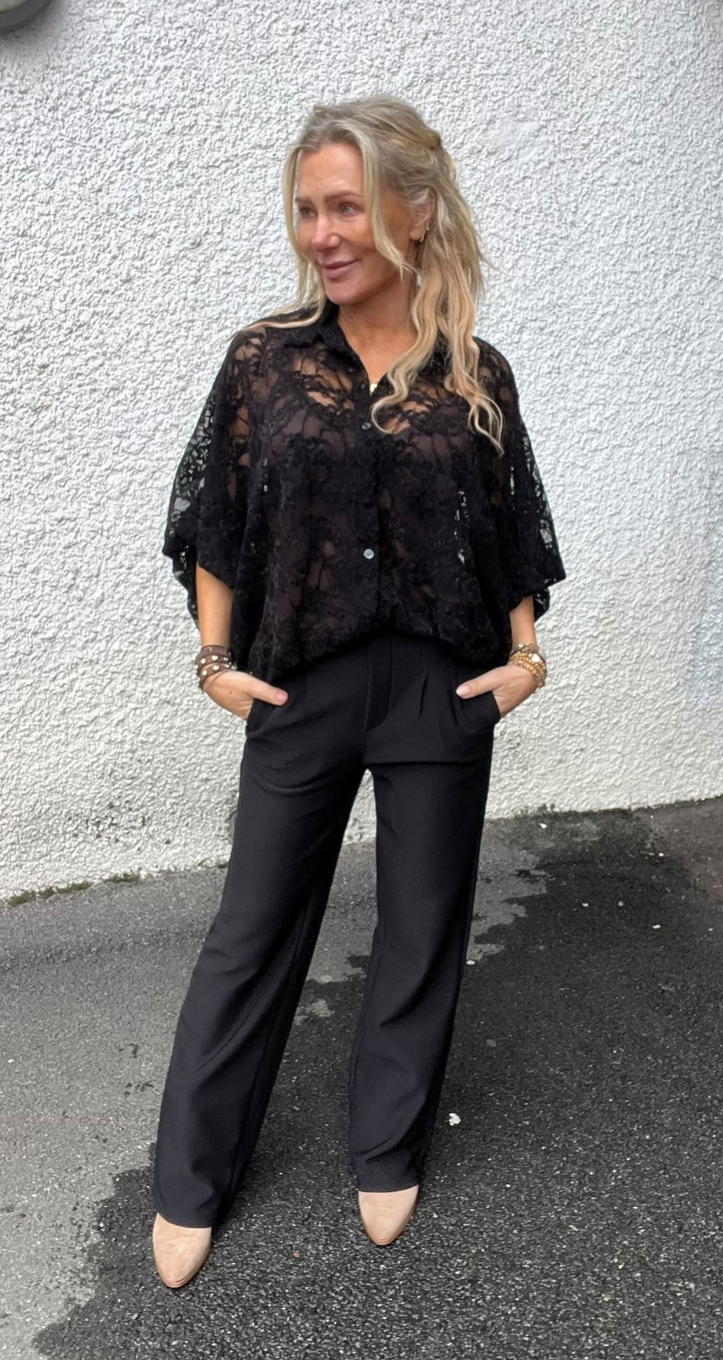 Blond kvinna står avslappnat med händerna i fickorna och ler åt sidan. Hon bär svart spetsblus med knappar, svarta byxor, beige skor och flera armband, mot en vit putsad vägg på asfalt.