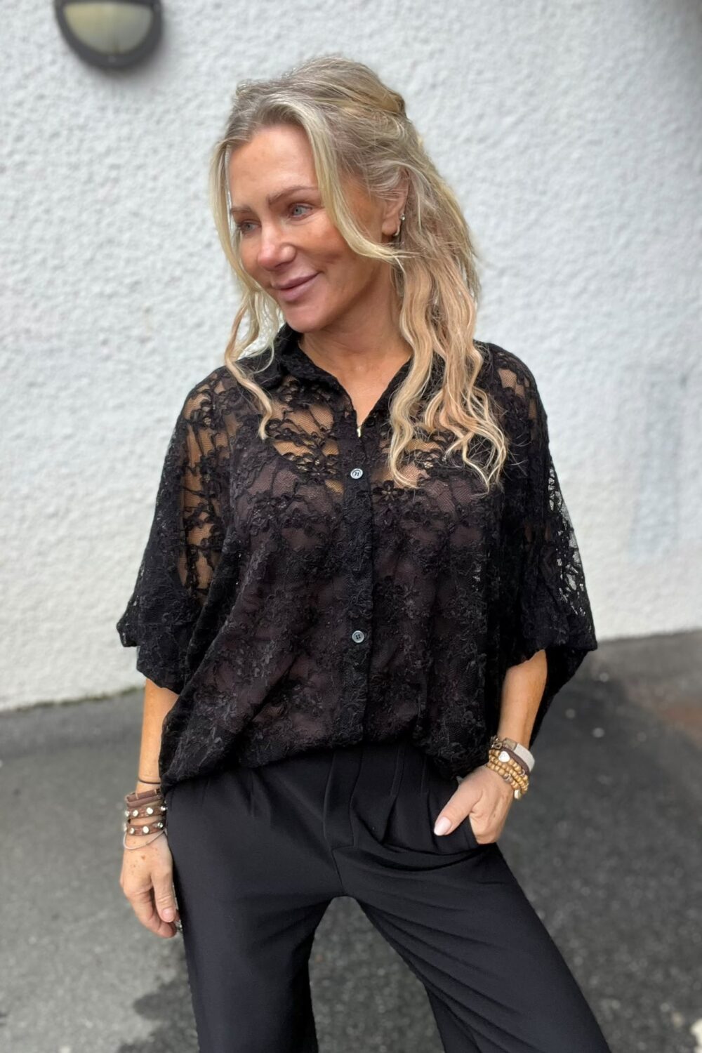 Leende kvinna med blont, lockigt hår står utomhus och tittar åt sidan, med ena handen i fickan. Hon bär svart spetsblus och svarta byxor samt flera armband, mot en ljus vägg med vägglampa.
