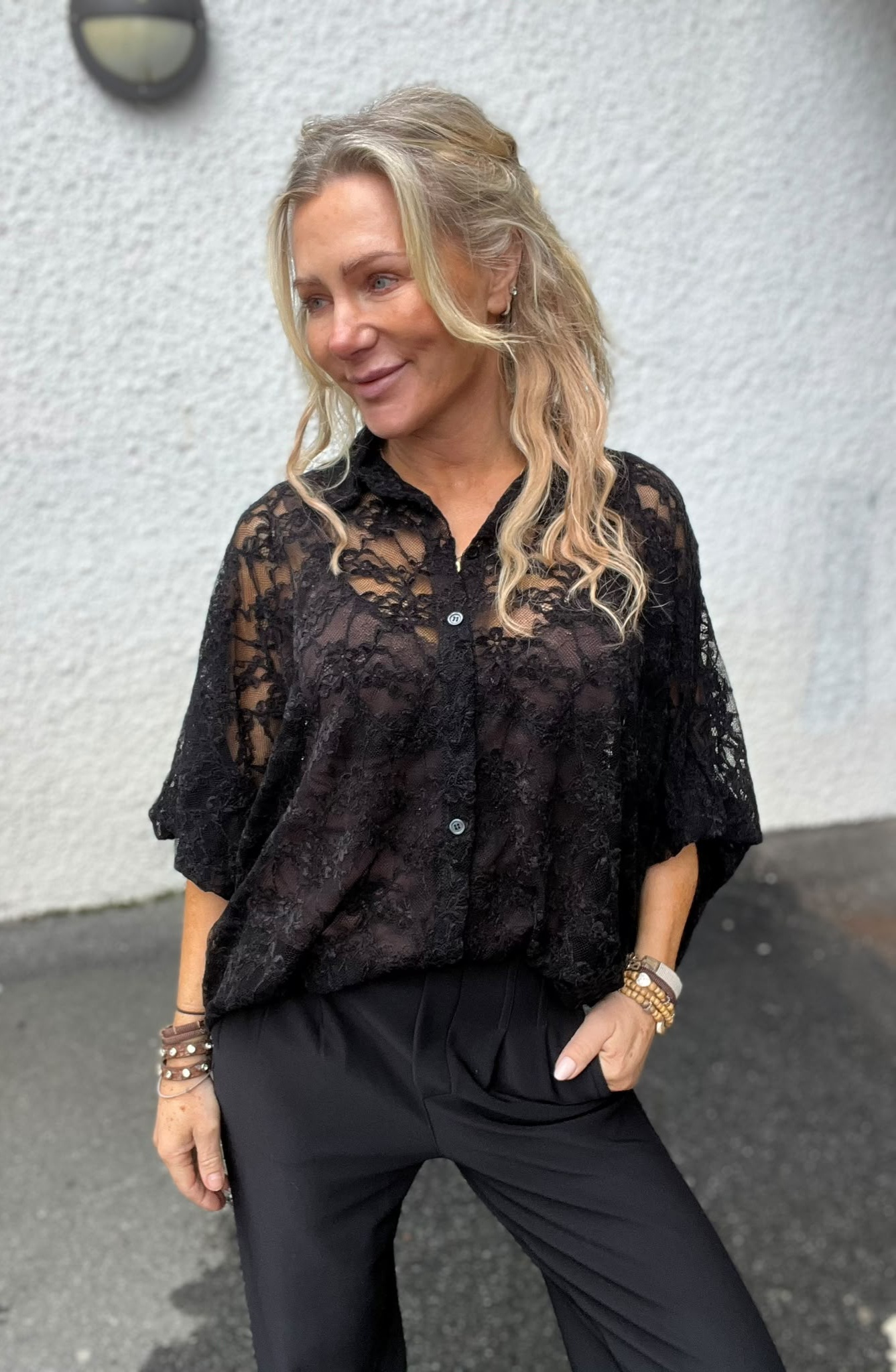 Leende kvinna med blont, lockigt hår står utomhus och tittar åt sidan, med ena handen i fickan. Hon bär svart spetsblus och svarta byxor samt flera armband, mot en ljus vägg med vägglampa.