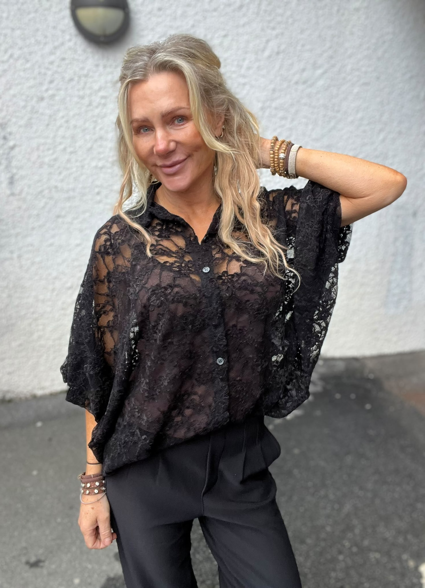 Leende kvinna med långt blont hår poserar med ena handen bakom huvudet. Hon bär en svart spetsblus och svarta byxor samt flera armband. Står utomhus framför en ljus vägg med en vägglampa.