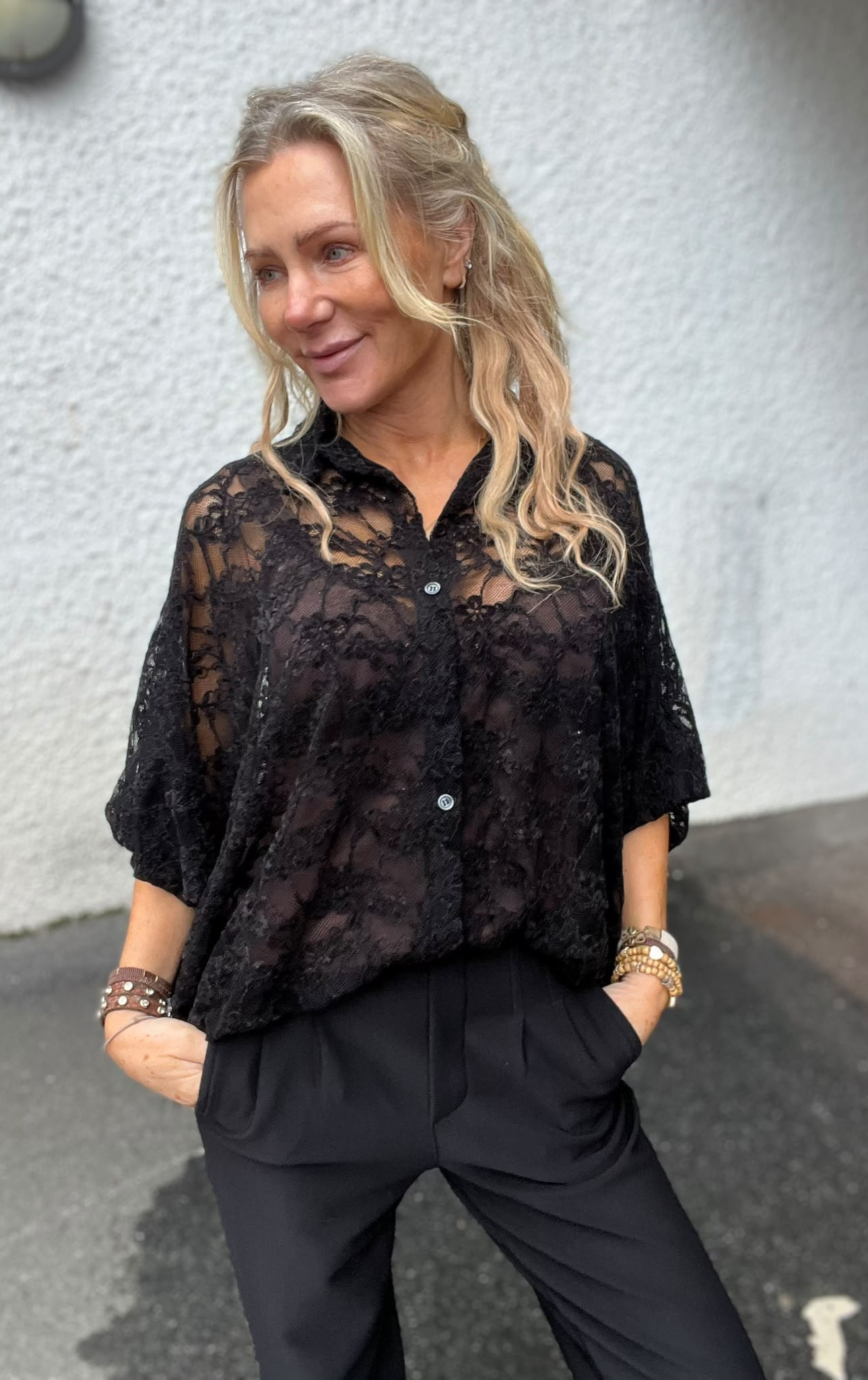 Blond kvinna står och ler lätt med händerna i fickorna, vänd åt sidan. Hon bär en svart, genomskinlig spetsblus med knappar och svarta byxor samt armband. Bakgrunden är en ljus vägg utomhus.