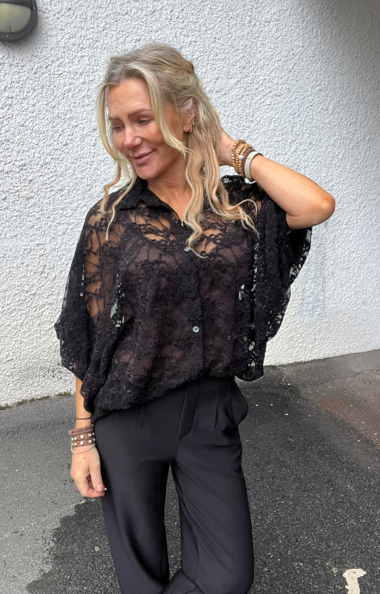 Blond kvinna står mot en ljusputsad vägg och ler medan hon lyfter ena handen till håret. Hon bär svart spetsblus och svarta byxor, med armband på handleden, på en asfaltbelagd yta utomhus.