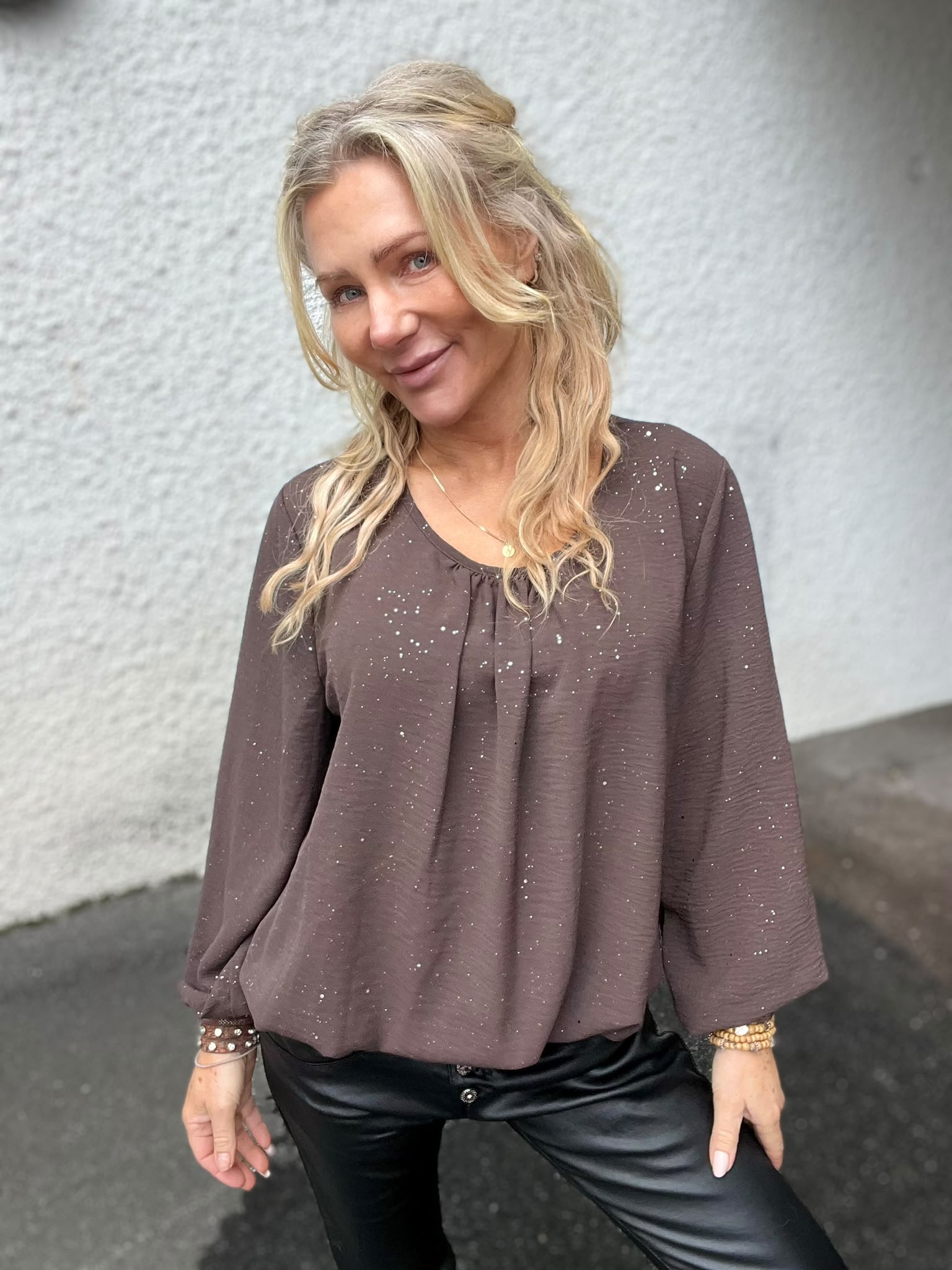 Blond kvinna står och ler mot kameran med huvudet lätt på sned, framför en ljus, grov vägg. Hon bär brun prickig blus, svarta läderbyxor, halsband och flera armband.