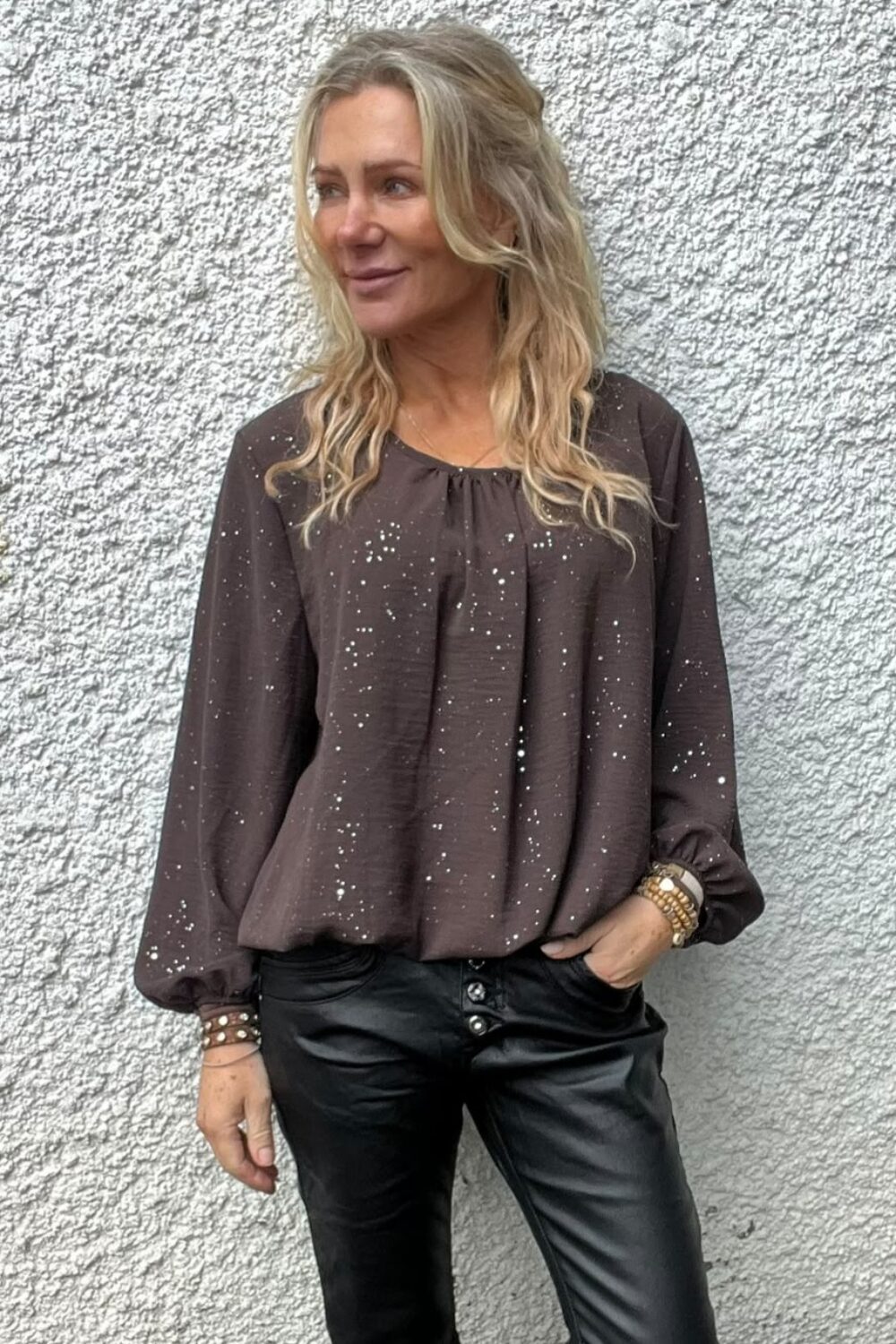 Blond kvinna står avslappnat mot en ljus, grovputsad vägg och tittar åt sidan. Hon har en mörkbrun prickig blus med långa ärmar, svarta skinnbyxor och armband, med ena handen i fickan.
