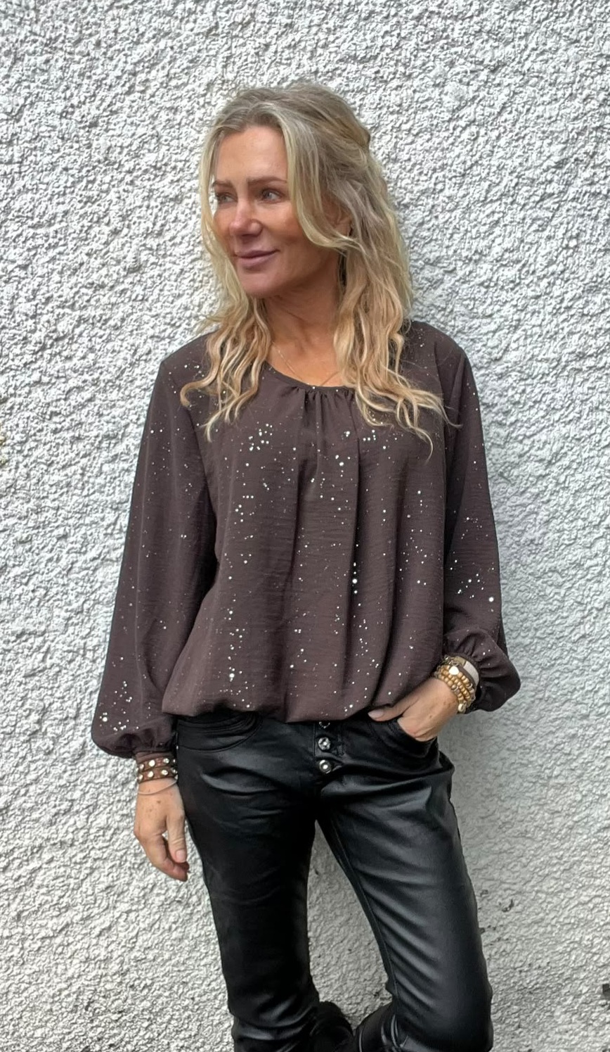 Blond kvinna står avslappnat mot en ljus, grovputsad vägg och tittar åt sidan. Hon har en mörkbrun prickig blus med långa ärmar, svarta skinnbyxor och armband, med ena handen i fickan.