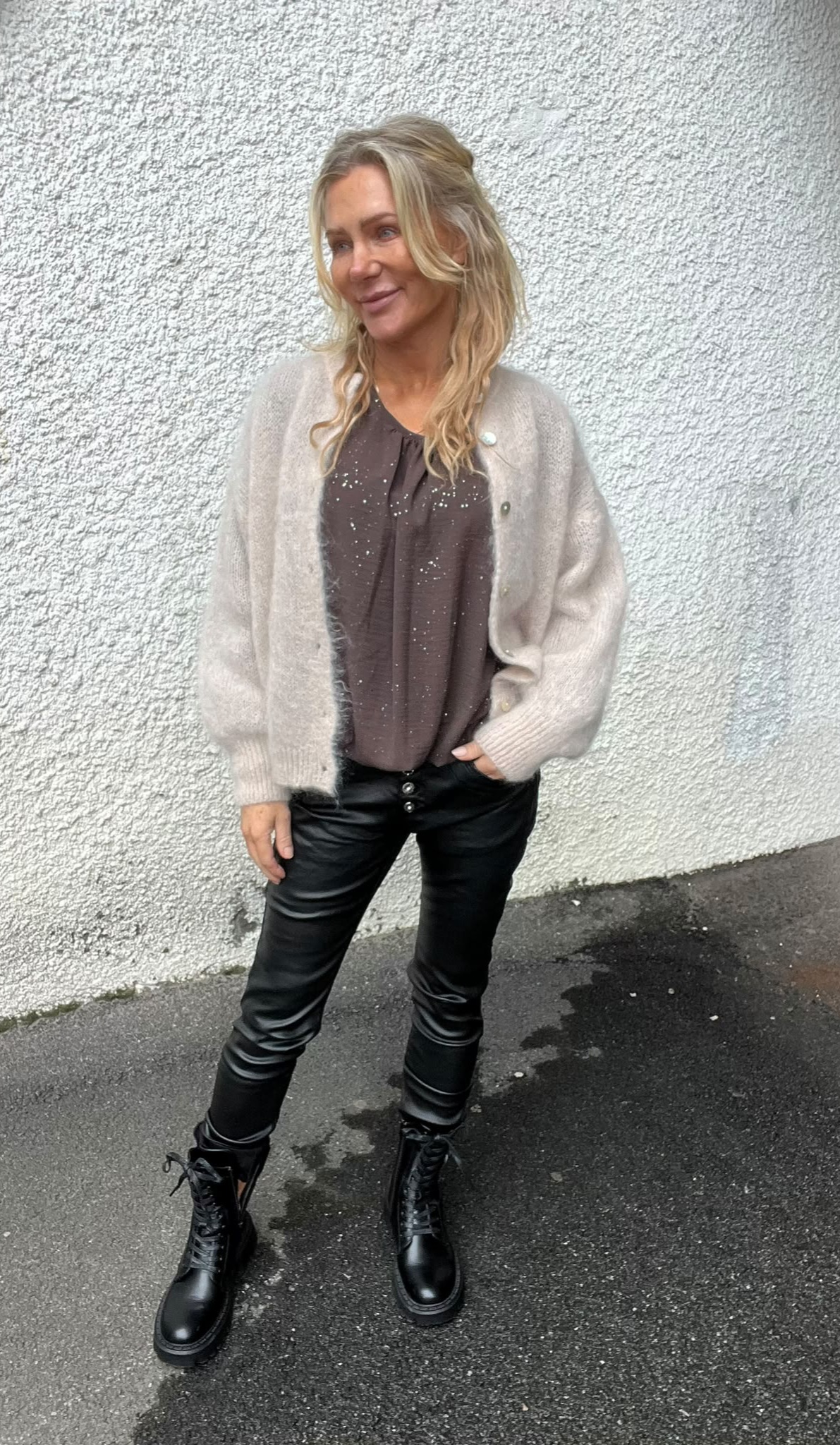 Blond kvinna står avslappnat och tittar åt sidan mot kameran, med ena handen i fickan. Hon bär ljus stickad kofta över glittrig mörk blus, svarta skinnbyxor och svarta kängor, framför vit grovputsad vägg.