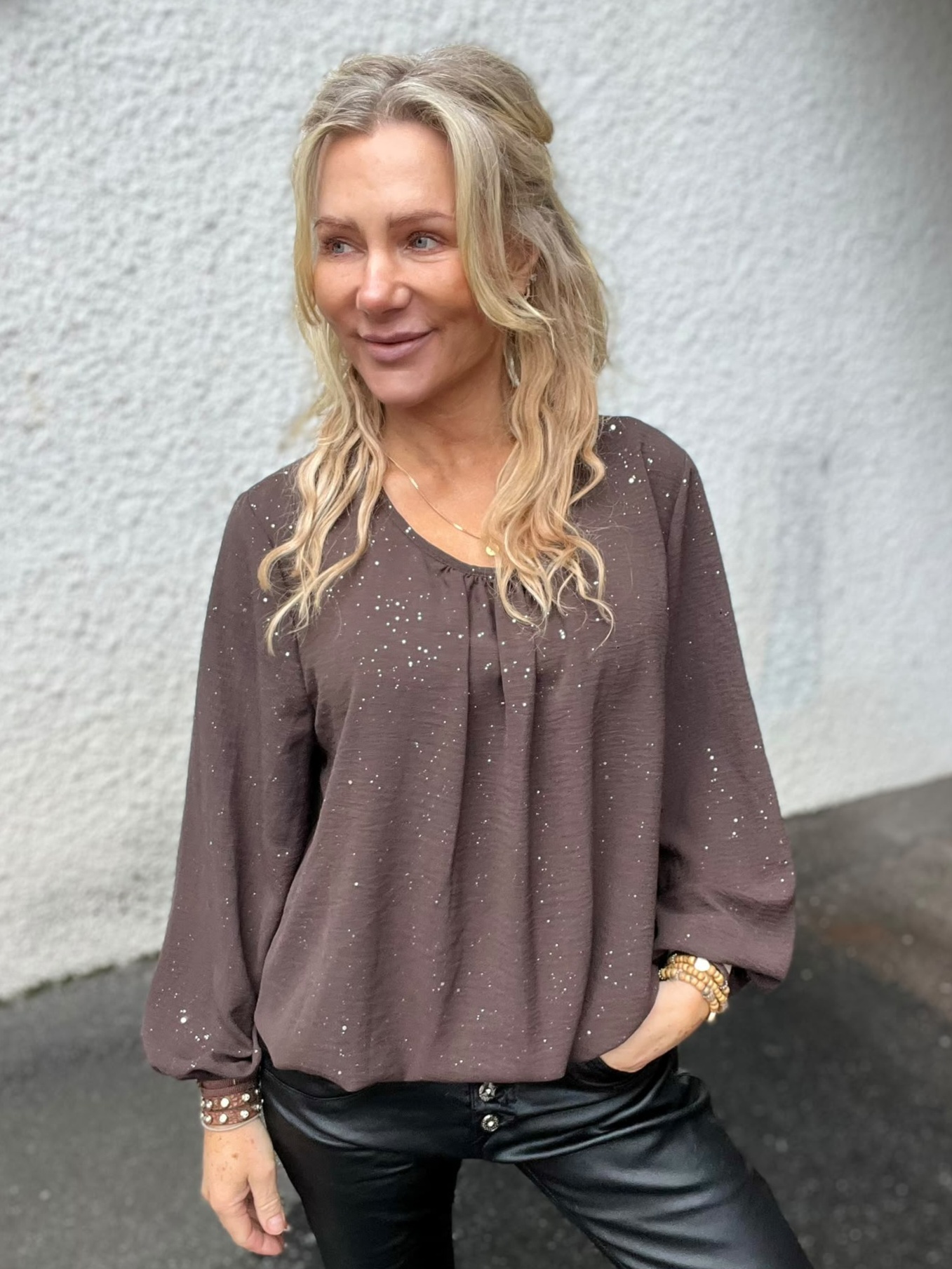 Blond kvinna står avslappnat med ena handen i fickan och ler medan hon tittar åt sidan. Hon bär brun, prickig blus och svarta skinnbyxor med armband, framför en ljus, grov vägg utomhus.