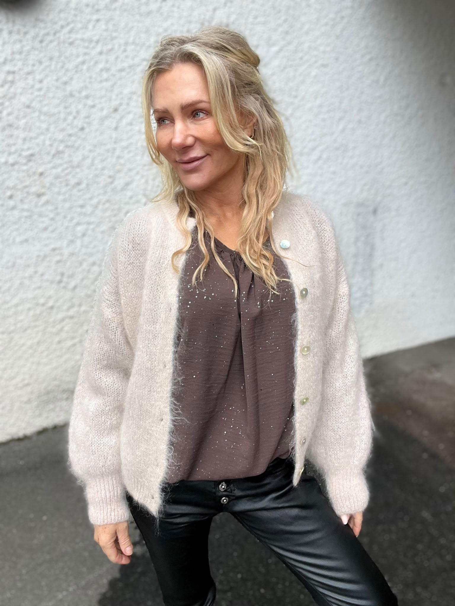 Blond kvinna står och ler lätt medan hon tittar åt vänster, klädd i beige stickad kofta, brun blus med små prickar och svarta skinnbyxor, framför en ljus, grov vägg utomhus.